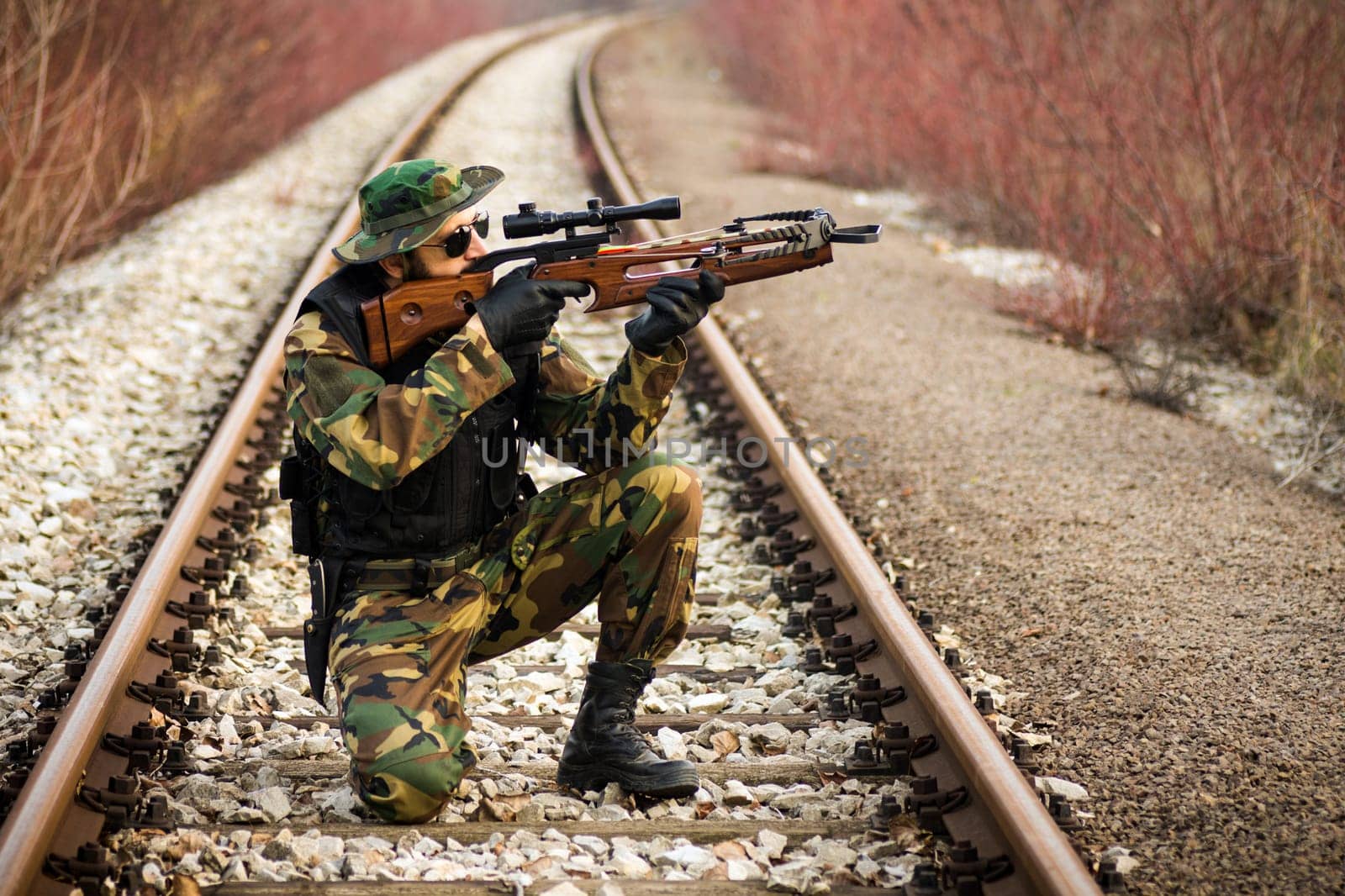 The soldier or hunter in military uniform is aiming with cross-bow on railway outdoors in nature.