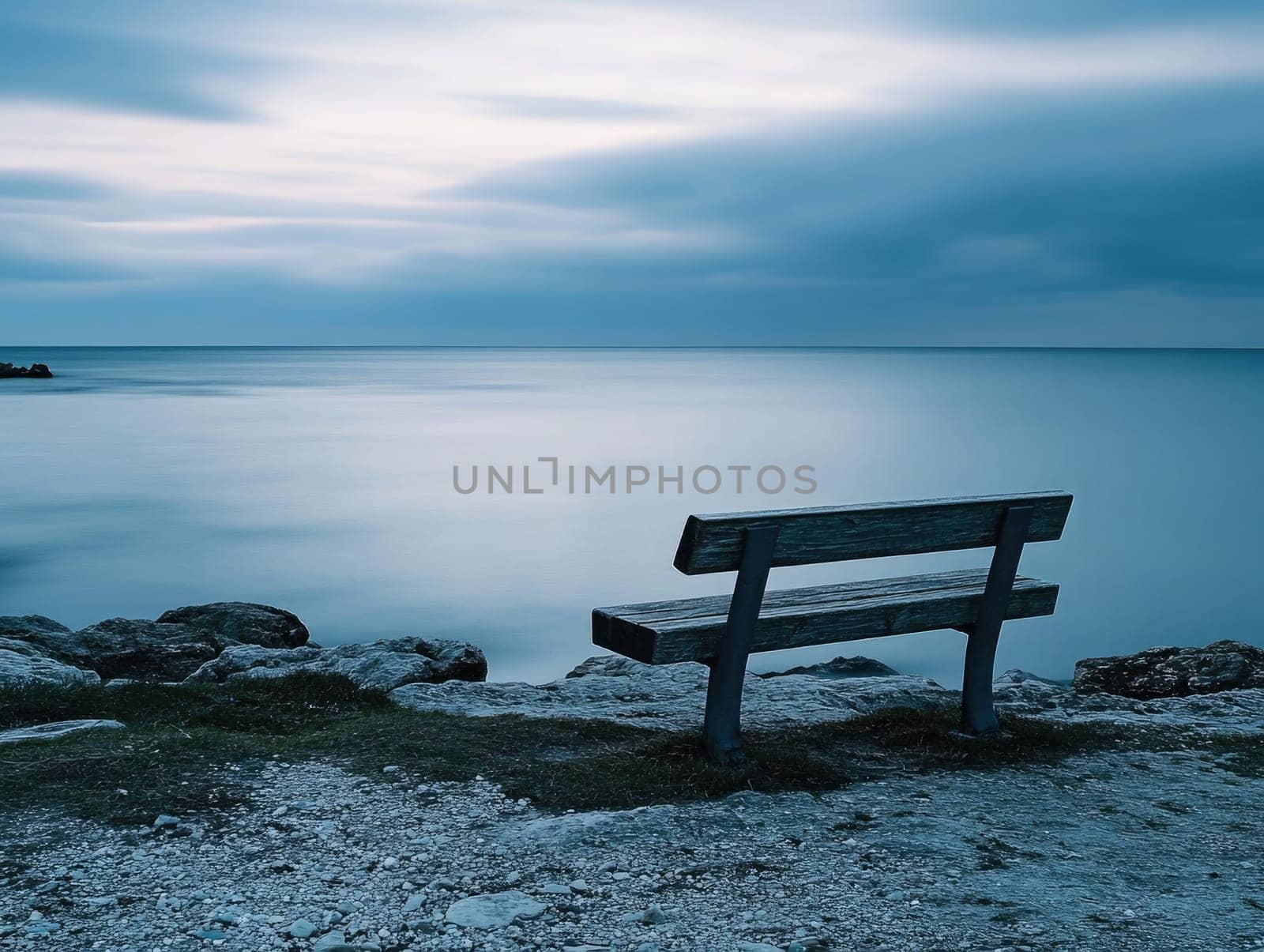 A wooden bench sits on the shore, facing the tranquil sea under a softly lit sky. The calm waters and gentle waves create a peaceful atmosphere during twilight.
