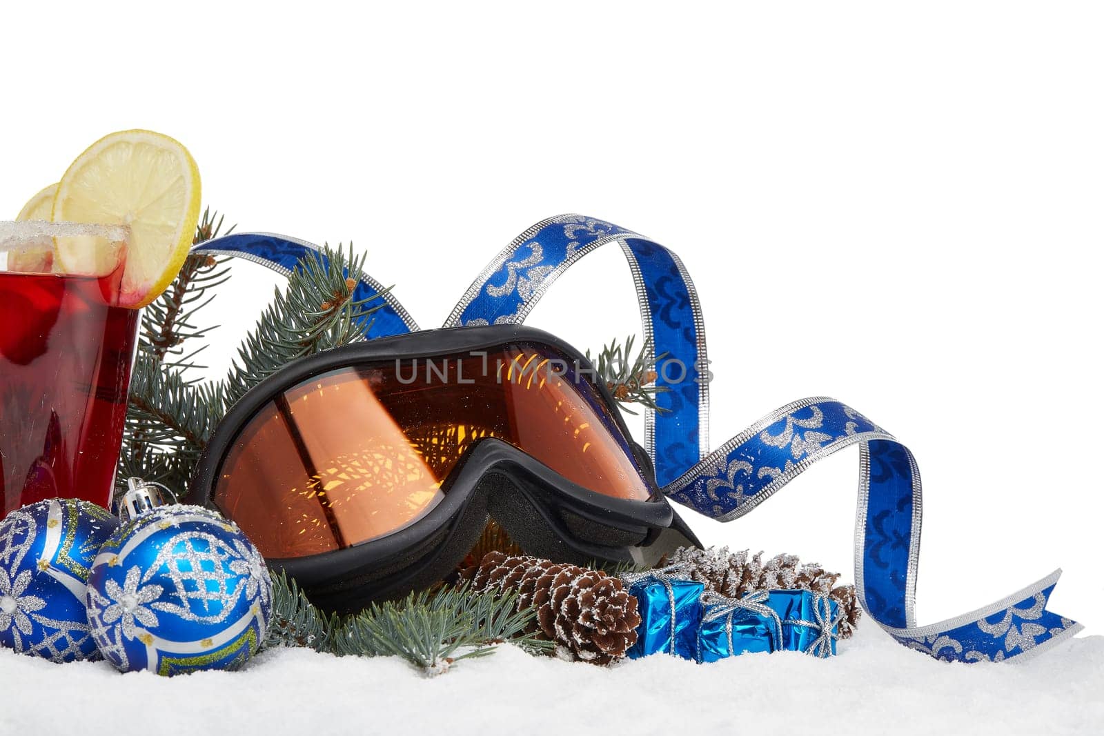 Winter holidays still life with fir tree branch, ski glasses, fruit tea and Christmas decorations lying on snow on a white background with copy space. New Year and Christmas. Winter vacation.