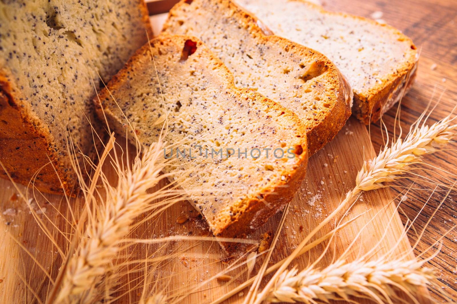 Close slice of fresh bread with poppy seeds and wheat ears on wooden background
