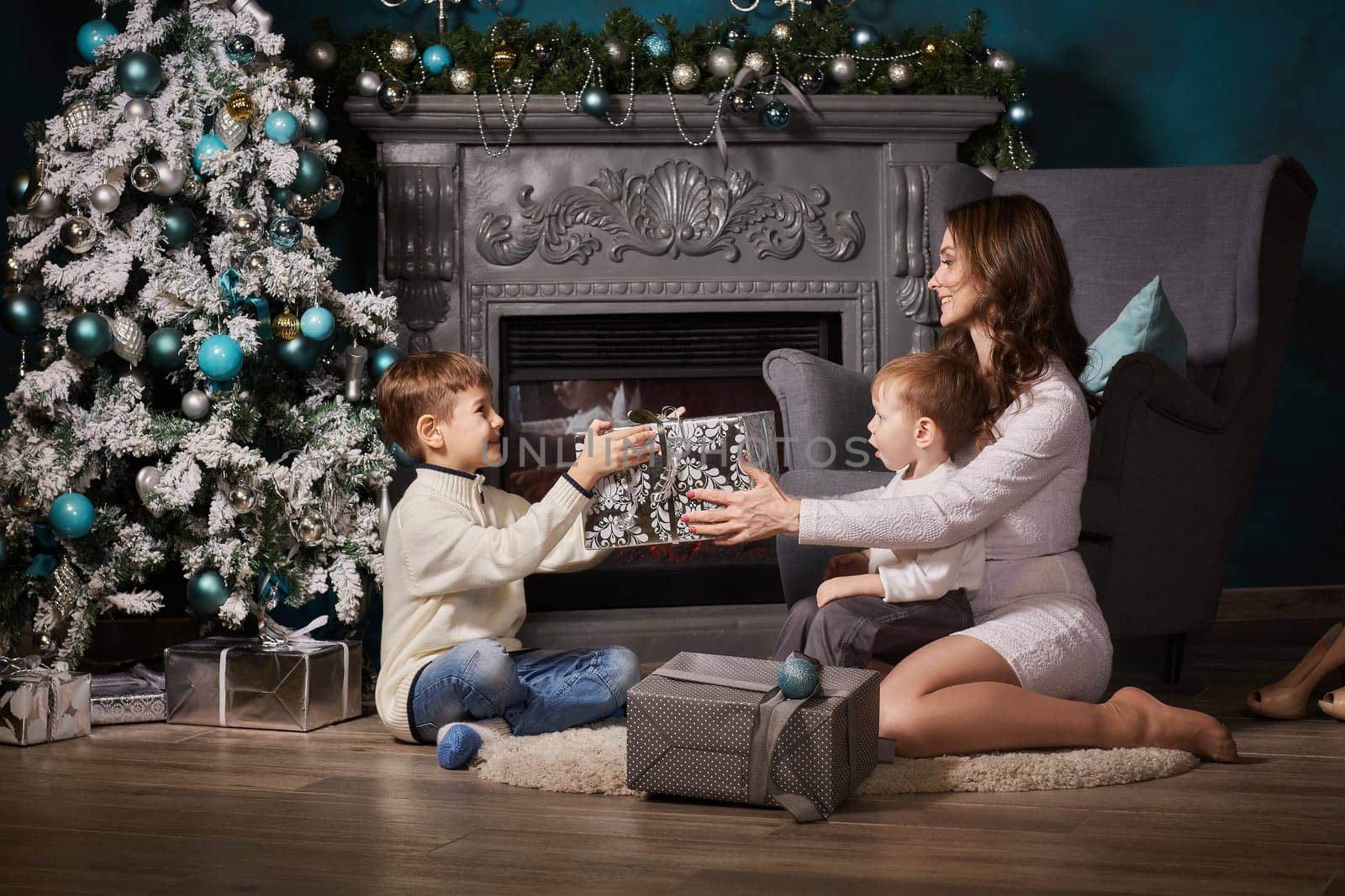 Happy family with gift boxes are sitting near Christmas tree at home. Merry Christmas and Happy New Year. Beautiful young mother with two sons.