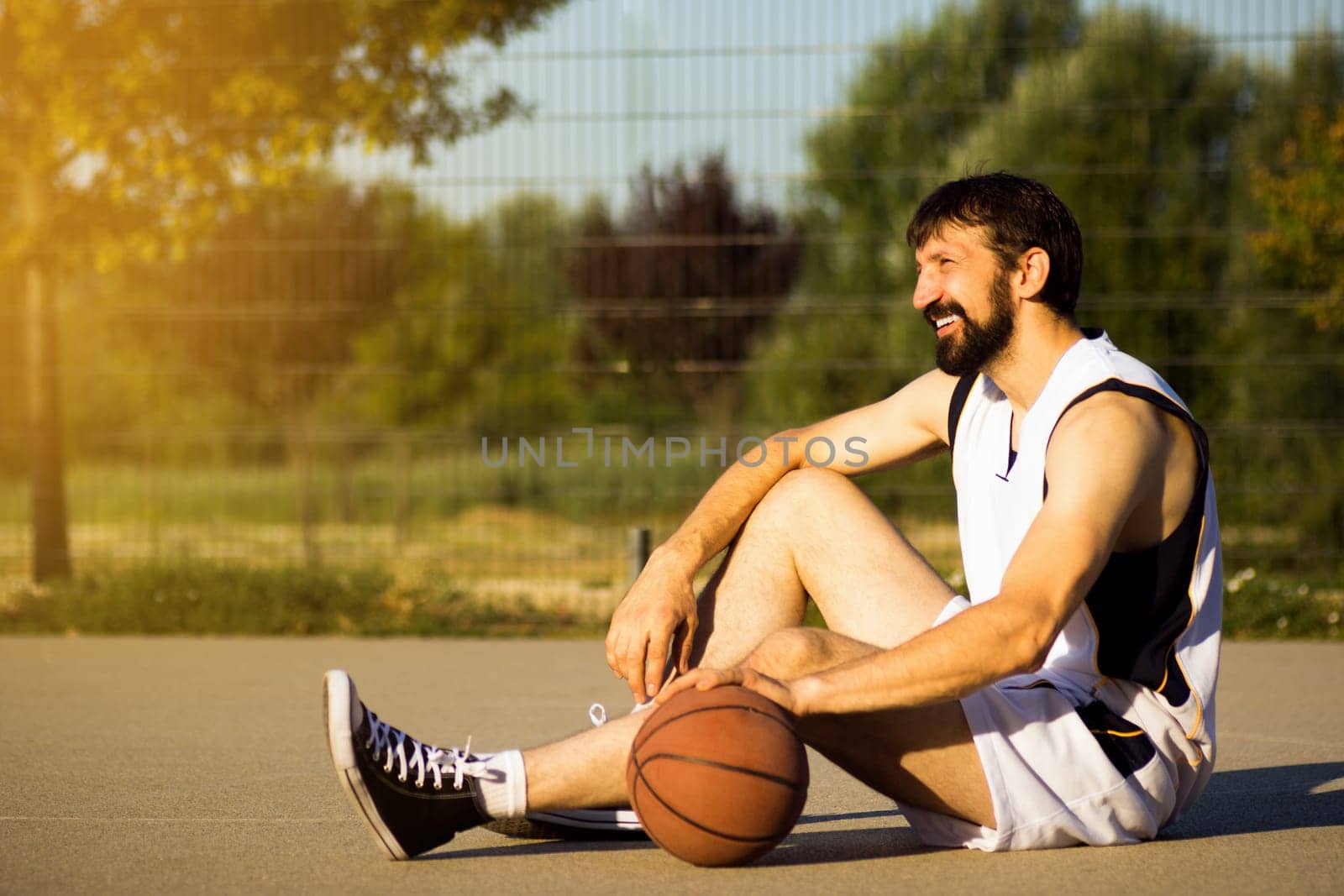 The smiling basketball player with basketball ball is sitting down on playground at the street outdoors and resting.