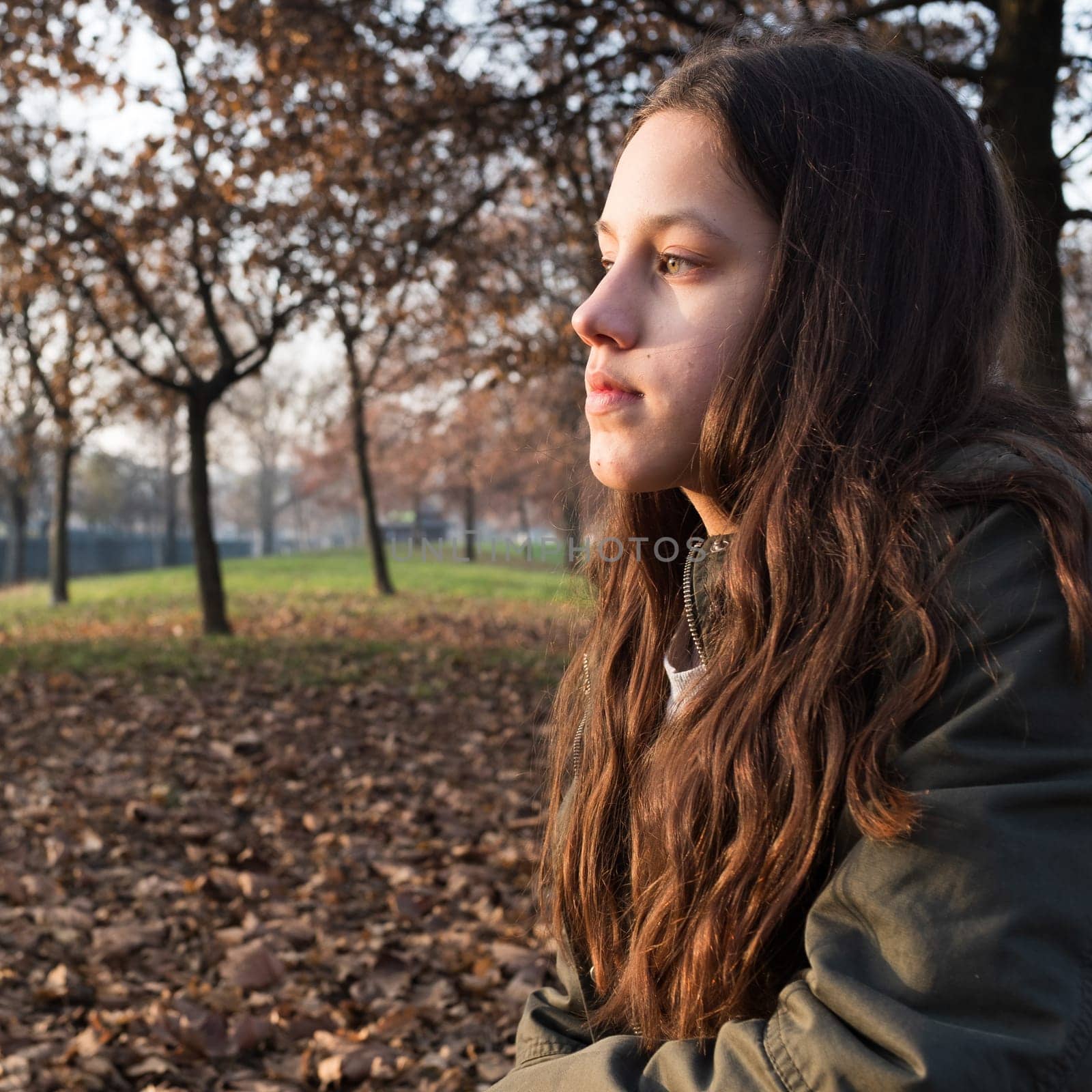 Thoughtful. Portrait of a serious young teen looking to the side in contemplation, an autumn background with foliage and trees. Attractive girl with cerulean eyes, long brown hair, green jacket. Square image