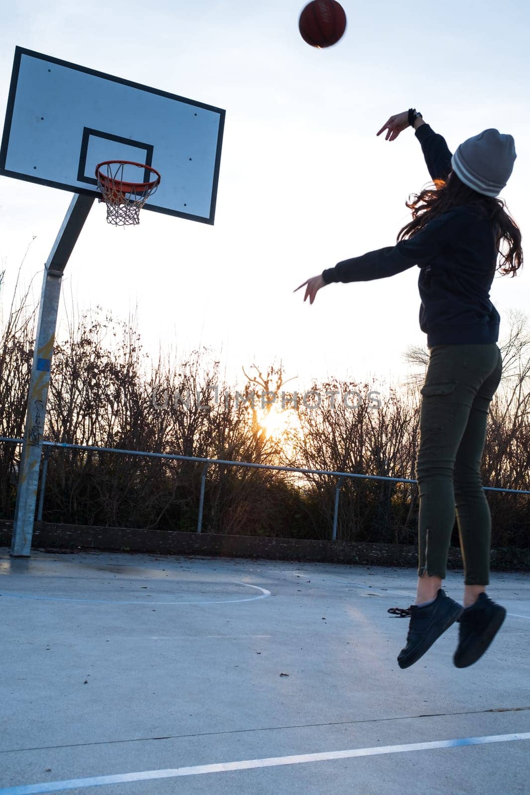 Young teen girl, mid air, shooting jump shot on basketball court, backlit at sunset, vertical composition, copy space