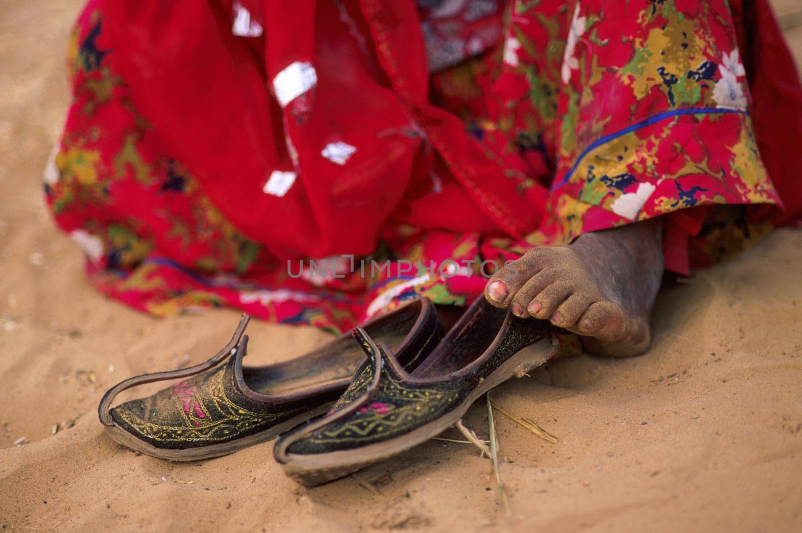 Low section of indian woman wearing a red sari and is sitting barefoot on sand after taking her shoes off, Pushkar, Rajasthan, India