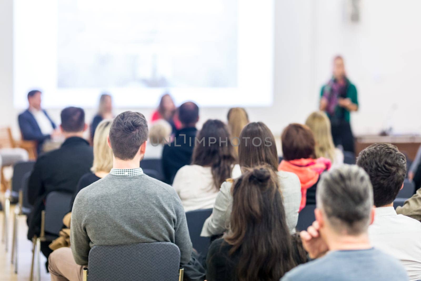 Business and entrepreneurship symposium. Female speaker giving a talk at business meeting. Audience in conference hall. Rear view of unrecognized participant in audience. Copy space on whitescreen.