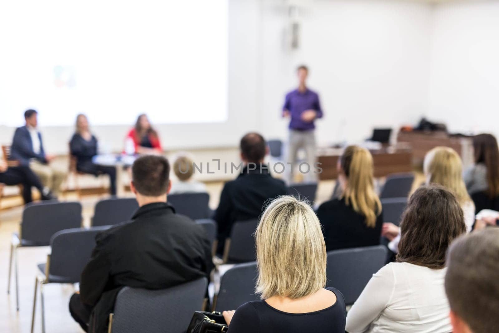 Business and entrepreneurship symposium. Speaker giving a talk at business meeting. Audience in the conference hall. Rear view of unrecognized participant in audience.