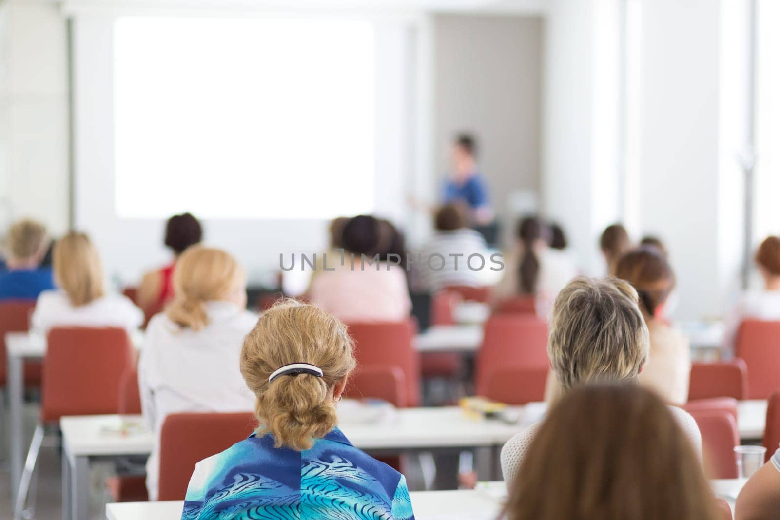 Female speaker giving presentation in lecture hall at university workshop. Audience in conference hall. Rear view of unrecognized participant in audience. Scientific conference event.