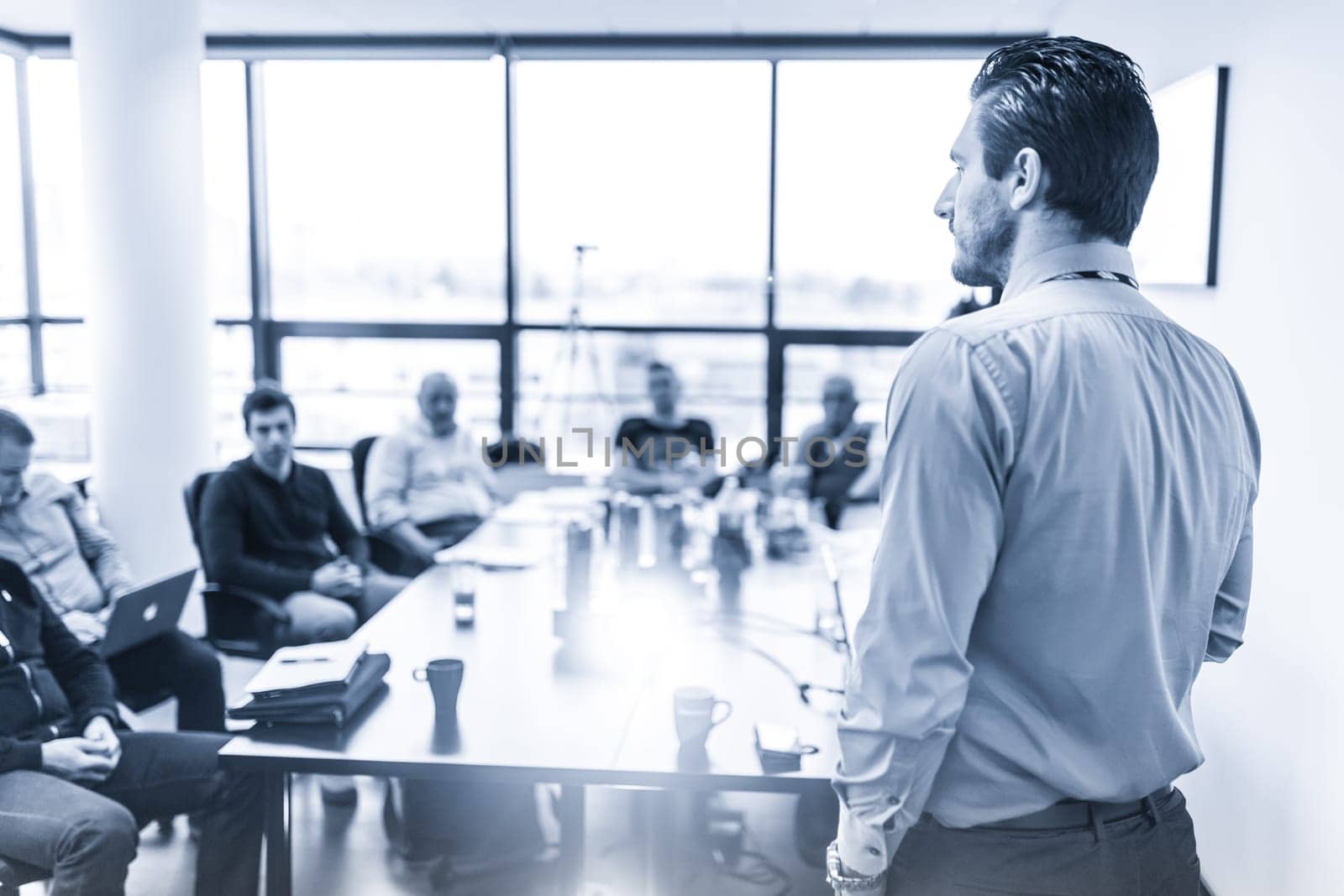 Business man making a presentation at office. Business executive delivering a presentation to his colleagues during meeting or in-house business training. Rear view. Blue toned.
