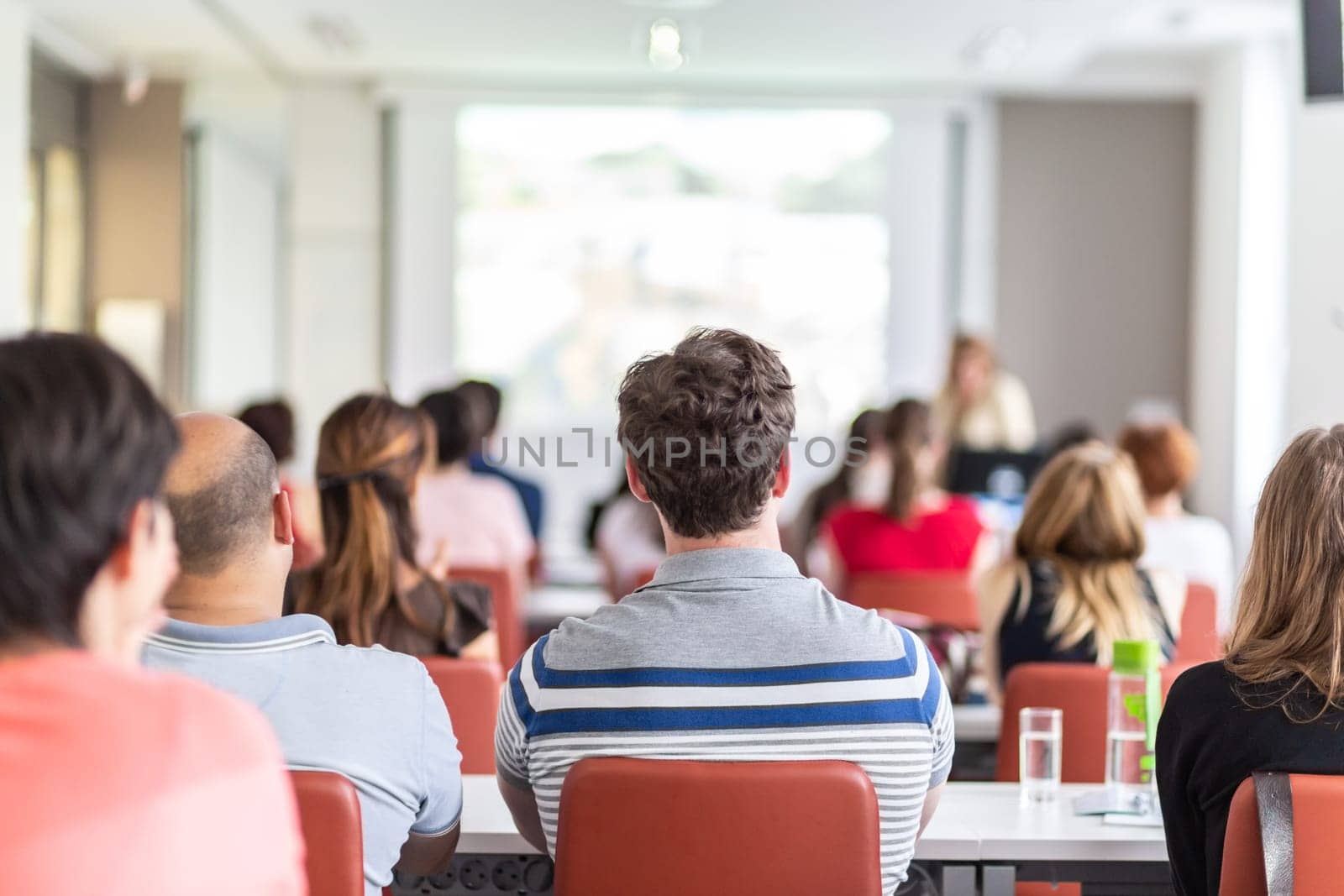 Female speaker giving presentation in lecture hall at university workshop. Audience in conference hall. Rear view of unrecognized participant in audience. Scientific conference event.