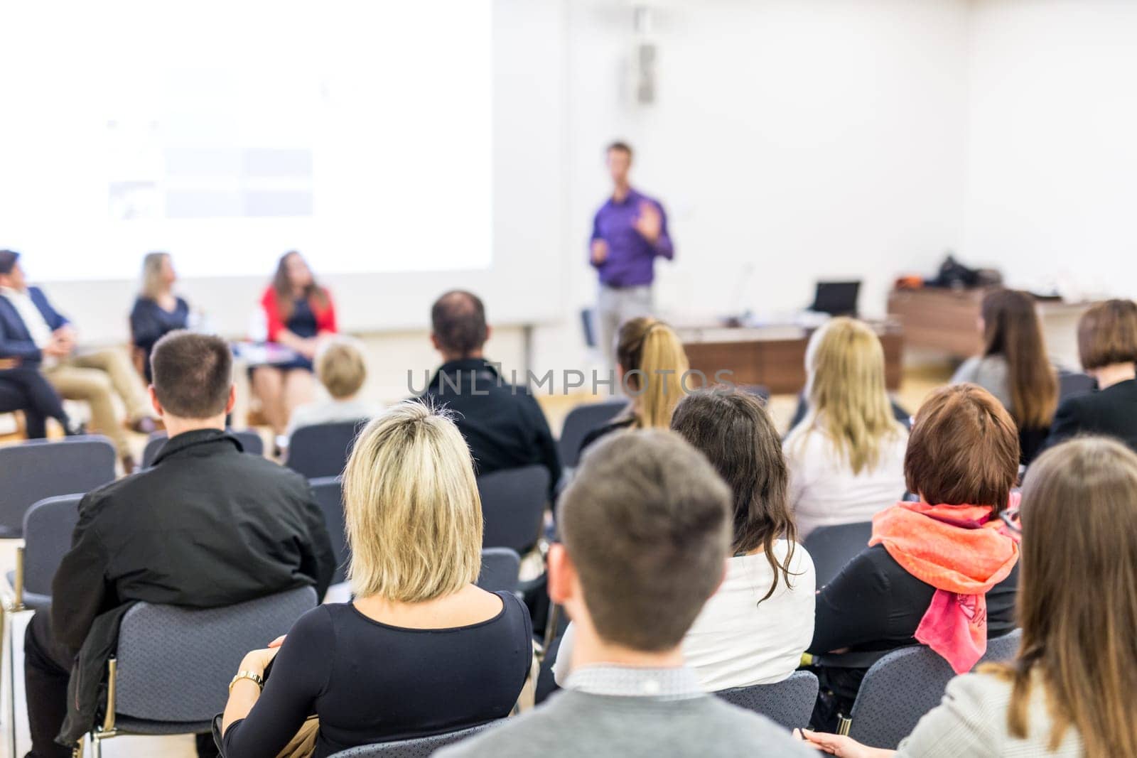 Business and entrepreneurship symposium. Speaker giving a talk at business meeting. Audience in the conference hall. Rear view of unrecognized participant in audience.