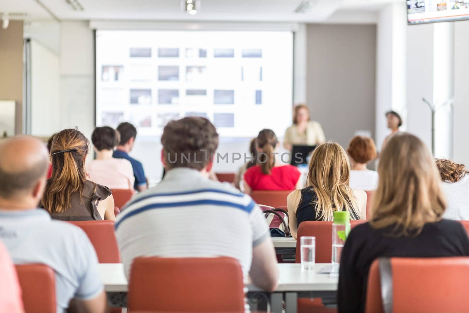 Female speaker giving presentation in lecture hall at university workshop. Audience in conference hall. Rear view of unrecognized participant in audience. Scientific conference event.