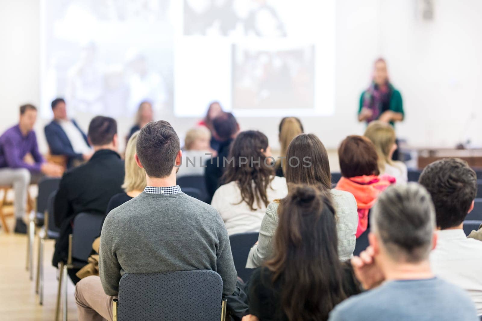 Business and entrepreneurship symposium. Female speaker giving a talk at business meeting. Audience in conference hall. Rear view of unrecognized participant in audience. Copy space on whitescreen.