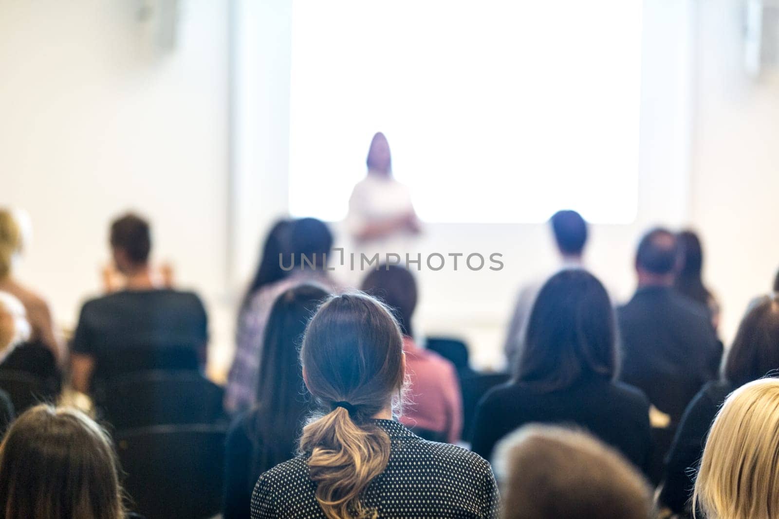 Business and entrepreneurship symposium. Female speaker giving a talk at business meeting. Audience in conference hall. Rear view of unrecognized participant in audience. Copy space on whitescreen.