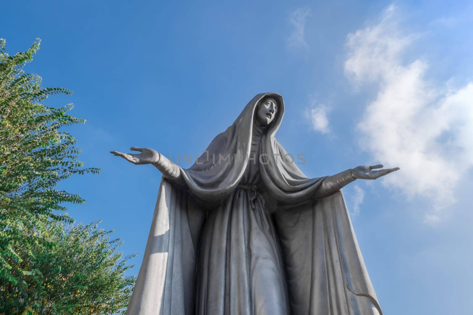 Statue of Virgin Mary in bronze, in a blue sky background with white clouds and tree, with outstretched arms.