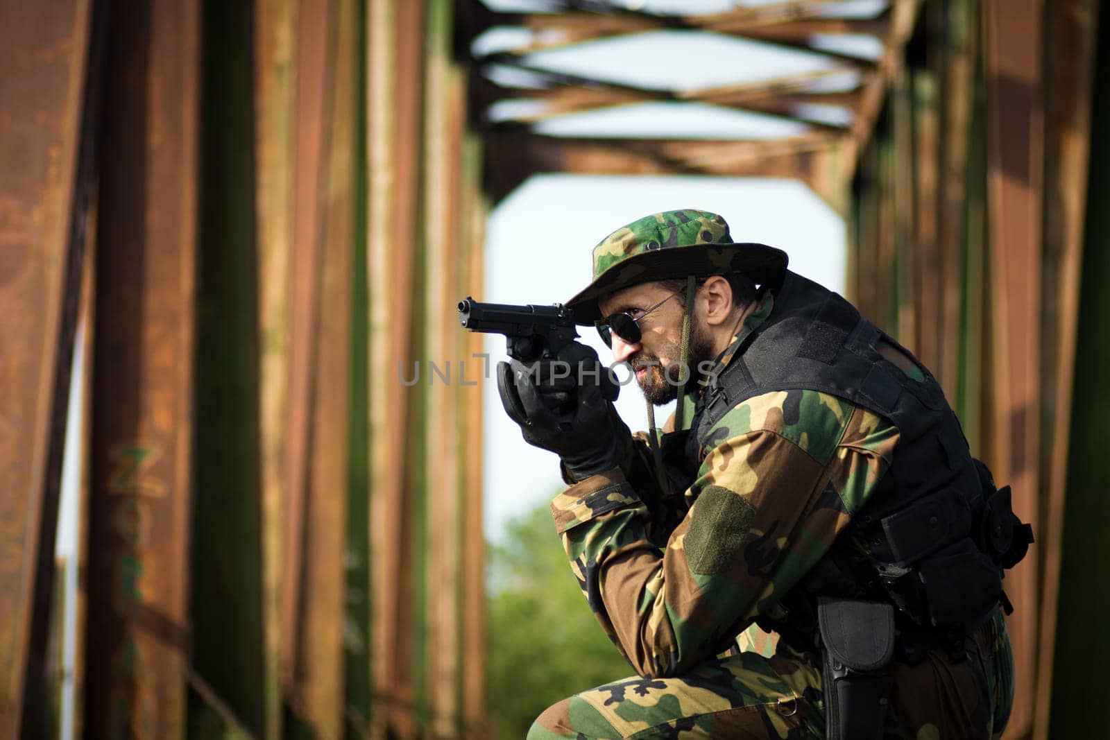The army soldier in military uniform is aiming with pistol outdoors on old bridge.