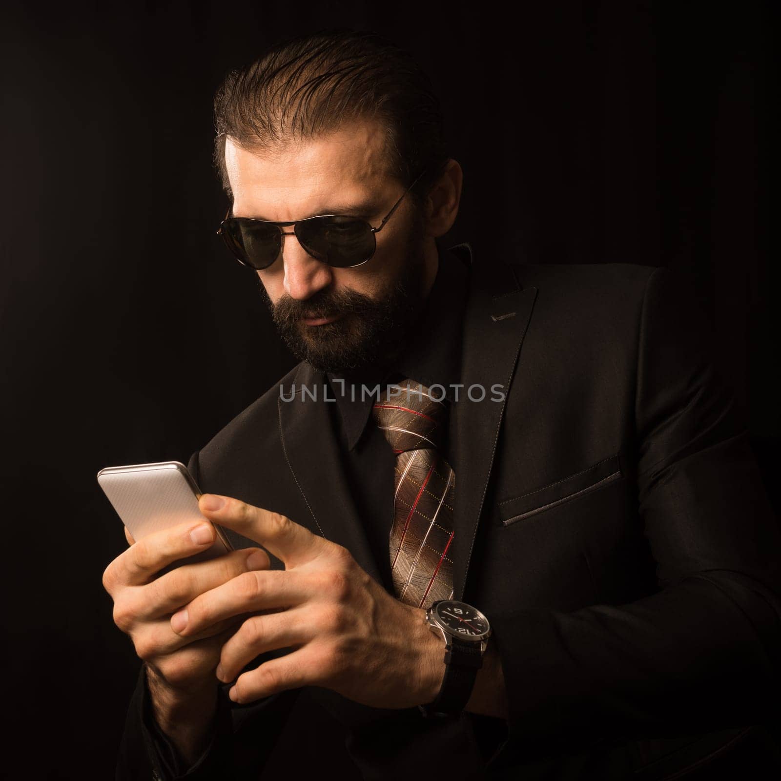 The elegant businessman in black suit is using phone on black background.