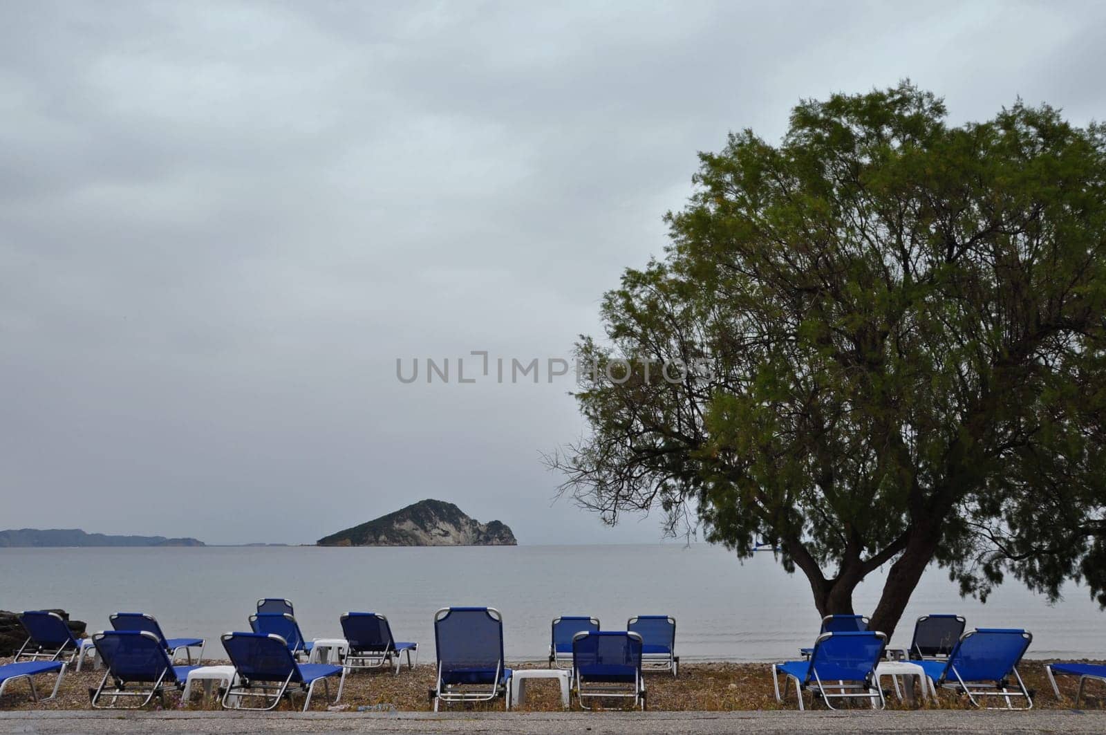 Lounge sunbed chairs with sea view tamarix tree and distant islet, Keri beach, Zakynthos Greece.