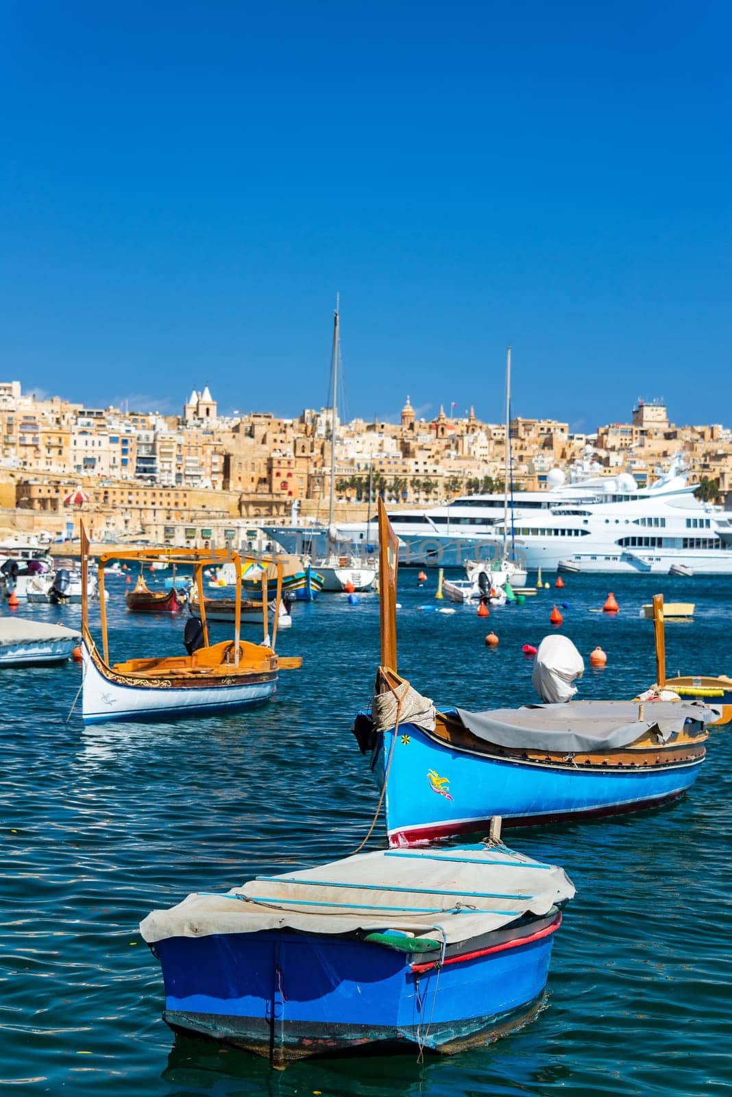 Traditional Maltese boats with superyachts in the background with Valletta visible in the background as seen from Senglea, Malta
