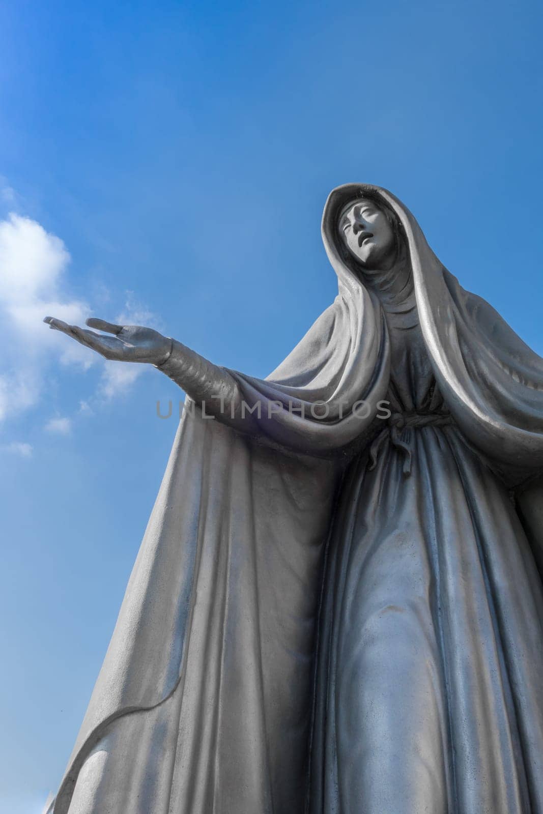 Statue of Virgin Mary in bronze, in a blue sky background with white clouds, with outstretched arms.