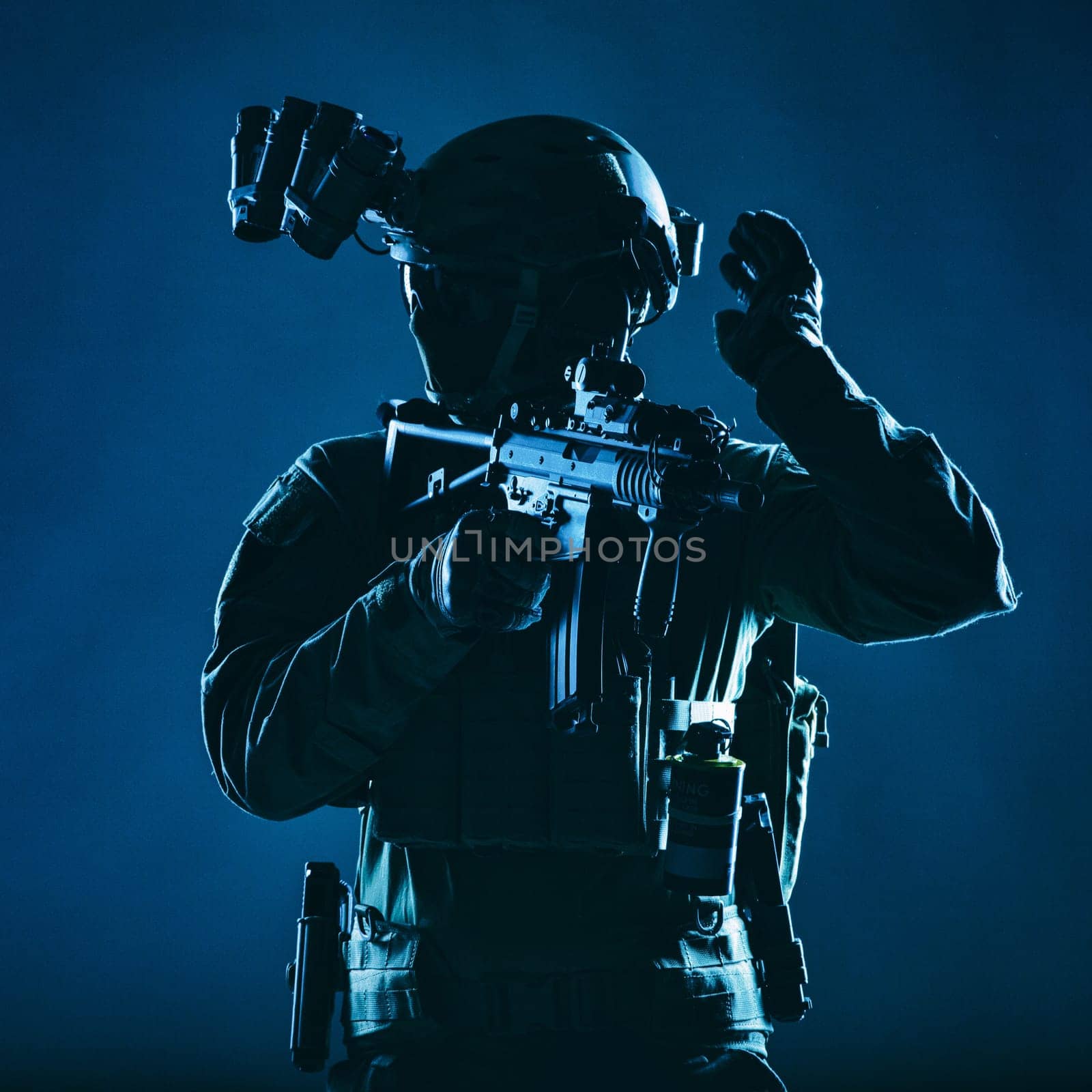 Special operations soldier, SWAT team fighter in mask and glasses, equipped night vision device, armed short barrel service rifle, looking back and showing freeze hand signal, low key studio shoot