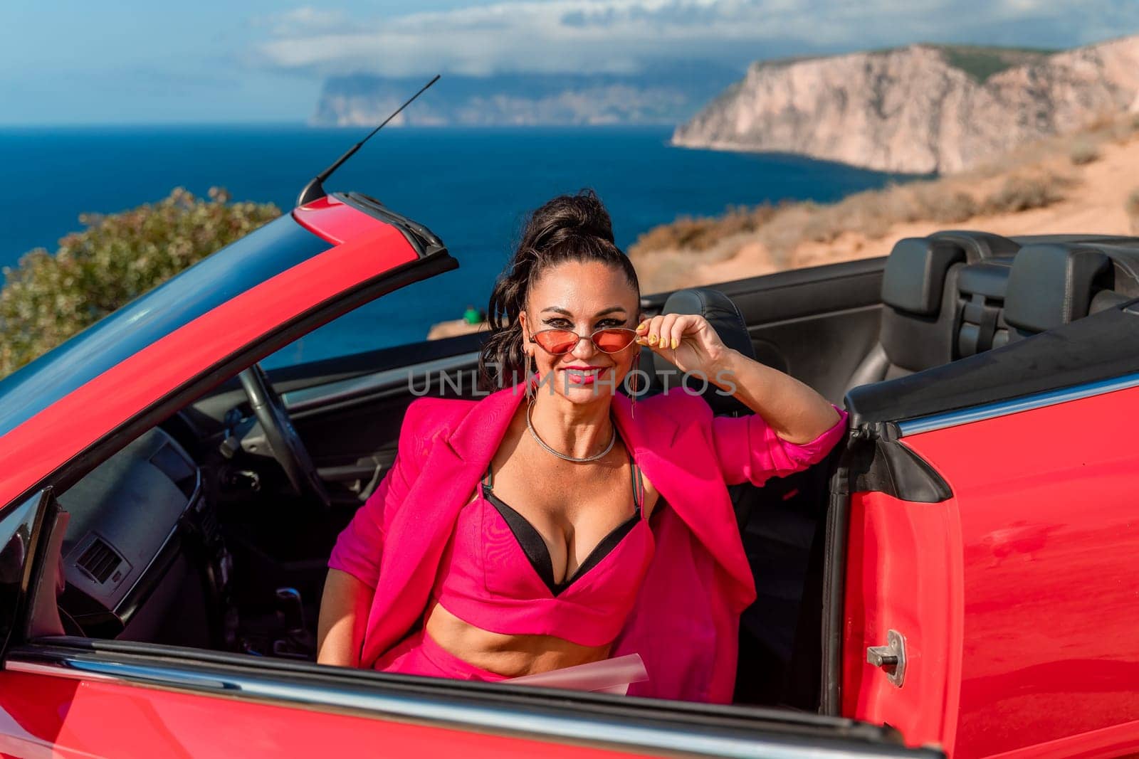 A woman in a pink suit is sitting in a red convertible. She is wearing sunglasses and a necklace