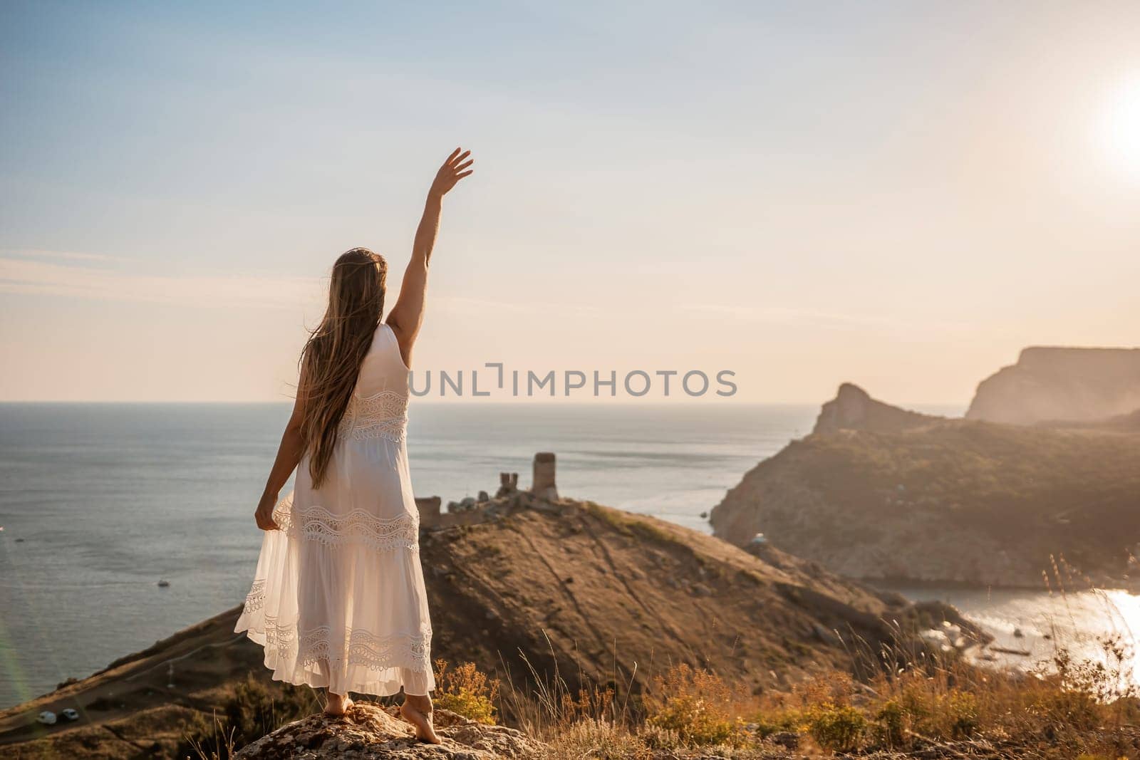 A woman in a white dress stands on a rocky hill overlooking the ocean. She is smiling and she is happy