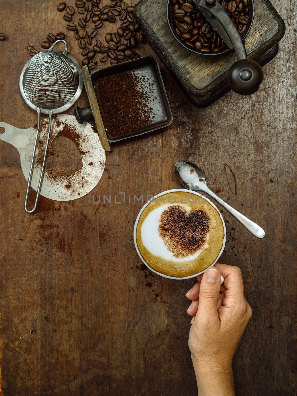 Photo of a messy rustic wooden table of coffee beans, grinder and a woman's hand holding a finished cup.
