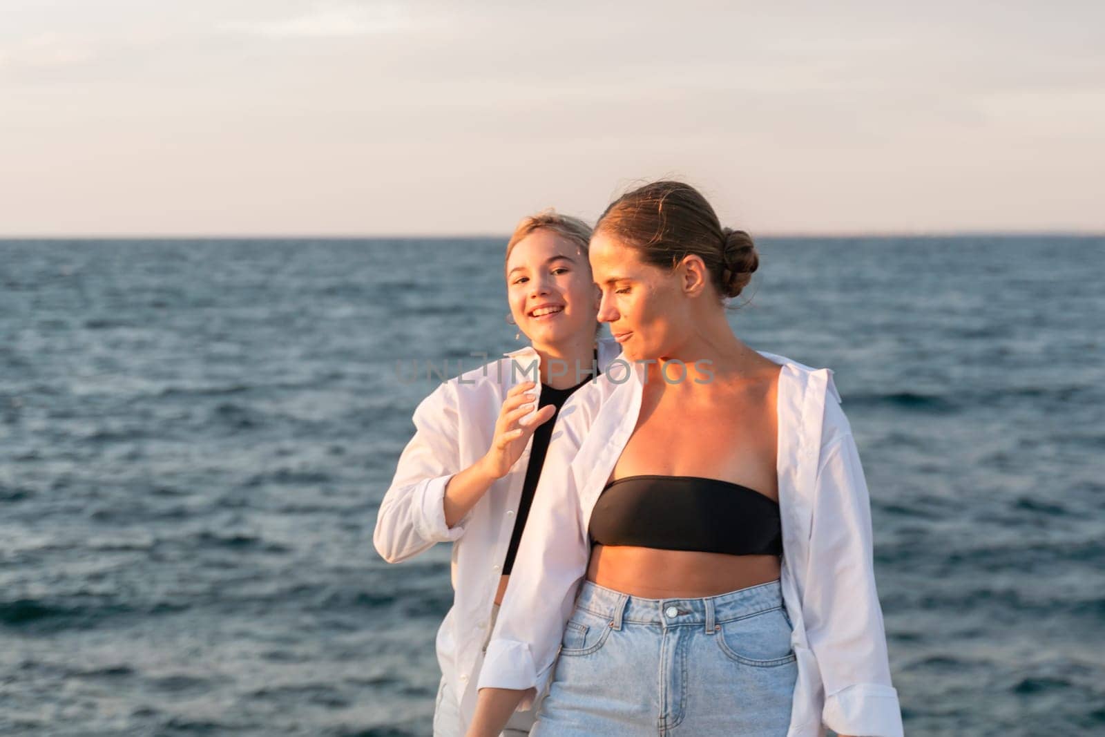 Two women, wearing white shirts, stand on a beach by the ocean. One woman wears a black bikini top and smiles at the camera. The other woman has a hand on her friend's shoulder and looks at her.