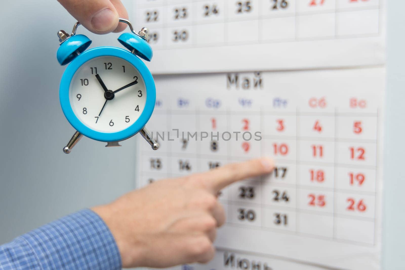 A hand holds a small alarm clock, in the background a hand points to long weekends and holidays on a wall calendar