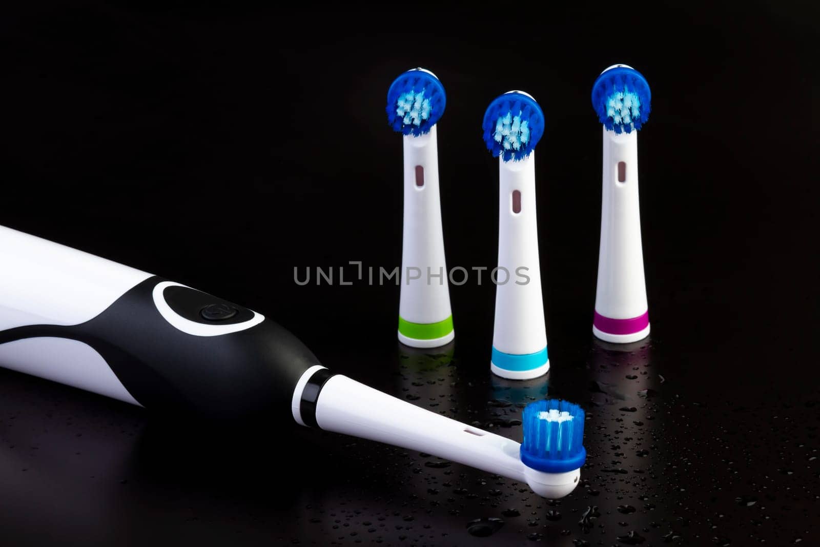 Electric rechargeable toothbrush with three toothbrush heads on black background with water drops. Focus on the power button.