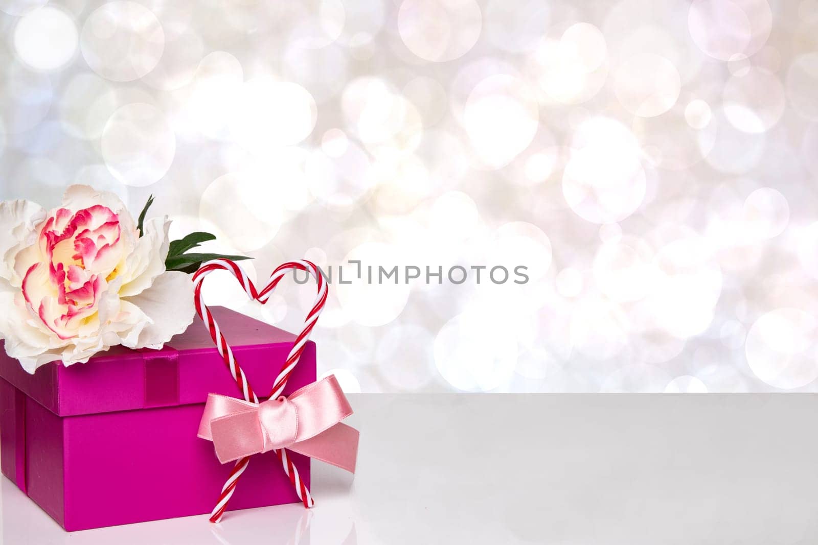 Happy Valentine or Mothers Day background. Red gift box with a beautiful white flower and red heart with bow ribbon on table over abstract bright bokeh backdrop. Space for your design.