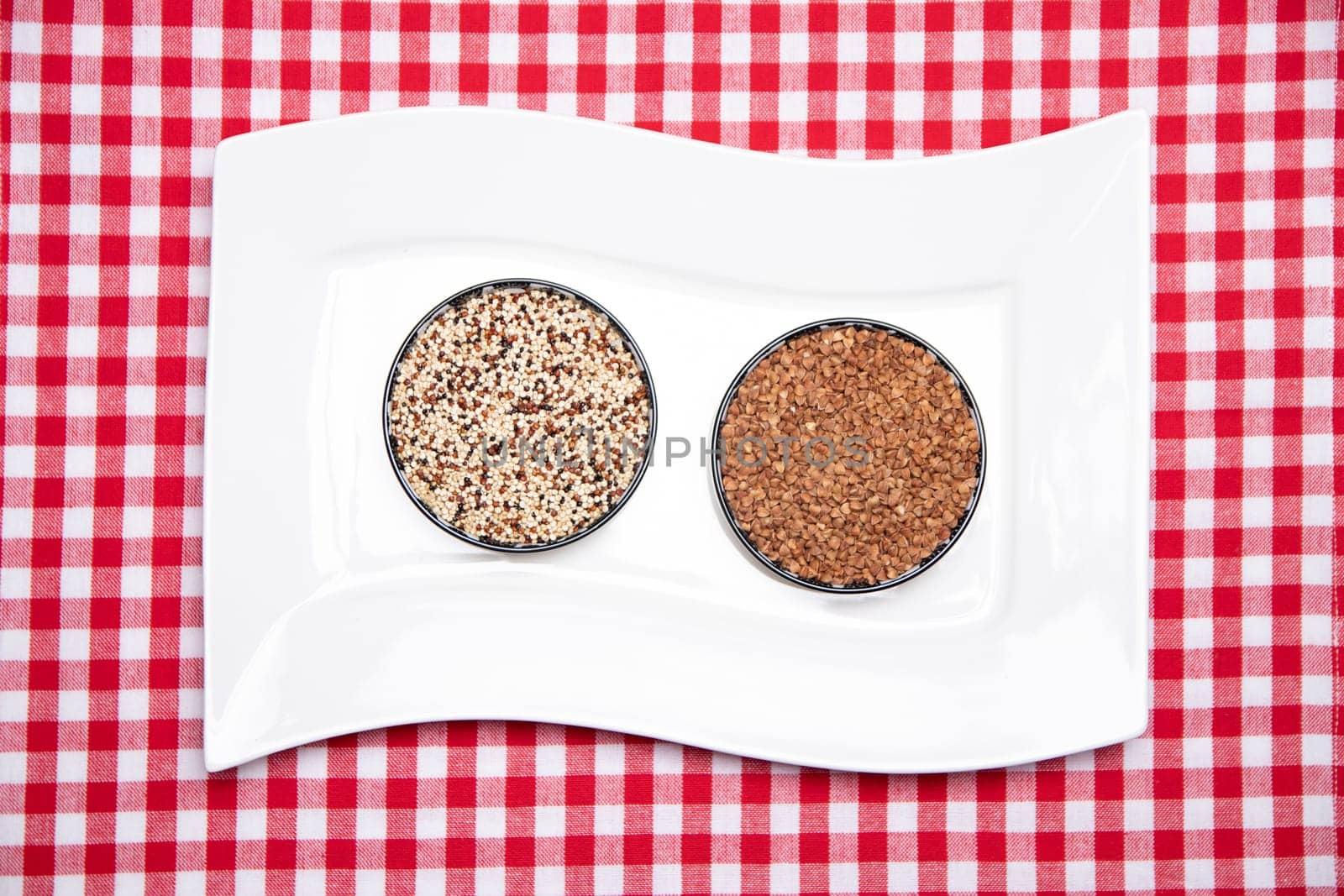 Vegetable food. A decorative white plate with two bowls with buckwheat and quinoa on a red checkered tablecloth. Healthy eating. Top view.
