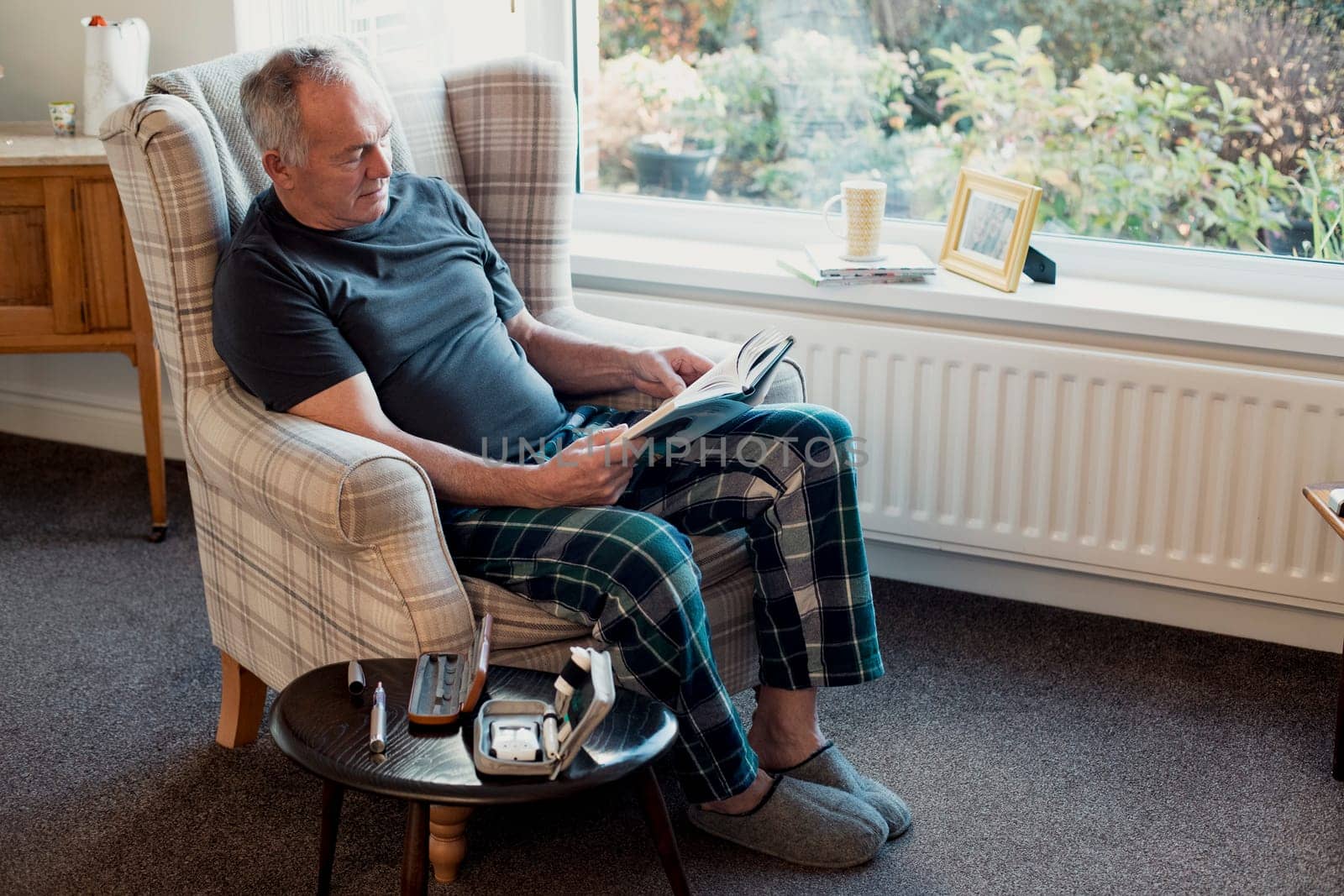 Senior diabetic man is relaxing in the living room of his home with a book. His blood glucose testing kit is on the table next to him.