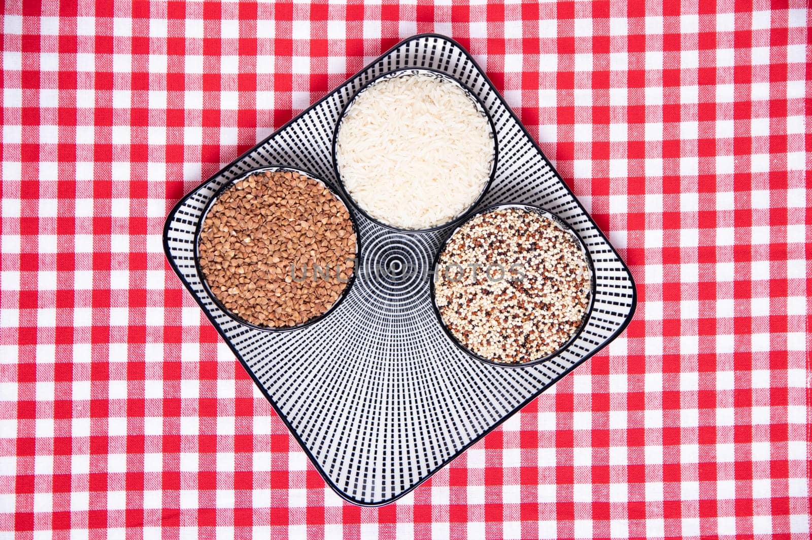 Vegetable food. A decorative plate with three bowls with buckwheat, rice and quinoa on a red checkered tablecloth. Healthy eating. Top view with space for design.