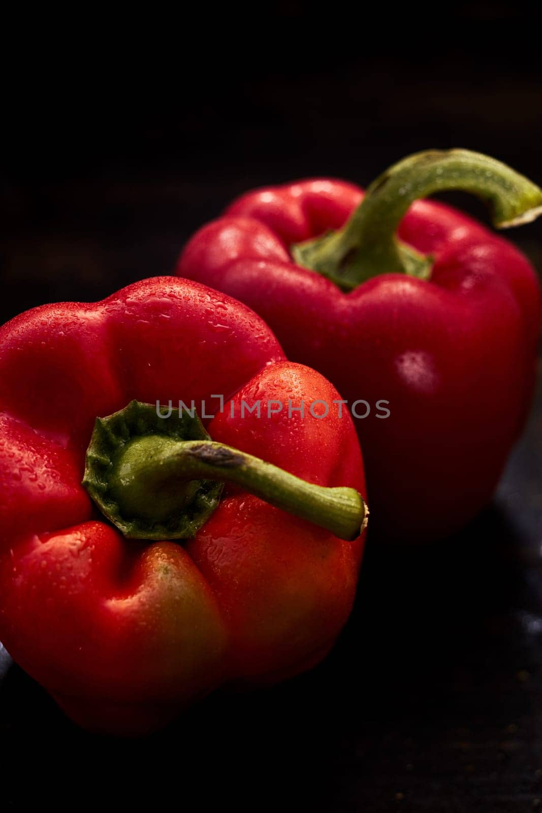Two fresh red healthy organic sweet peppers with water droplets on the skin and green stalks viewed close up on a dark background