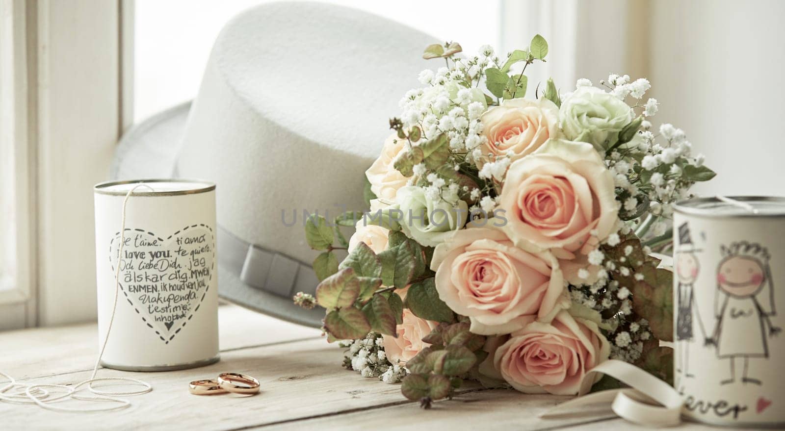 Bright composition for marriage day with wedding rings, bridal bouquet of pink roses, brooms white hat and cans with decorations and wishes, sitting on bright window sill