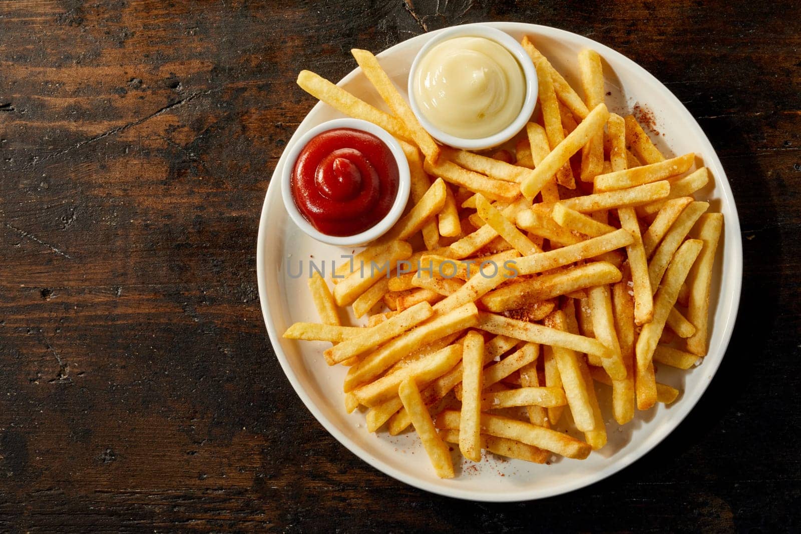 Plate of french fries potatoes served with ketchup and mayonnaise sauces in small bowls, viewed from above on dark wooden background with copy space