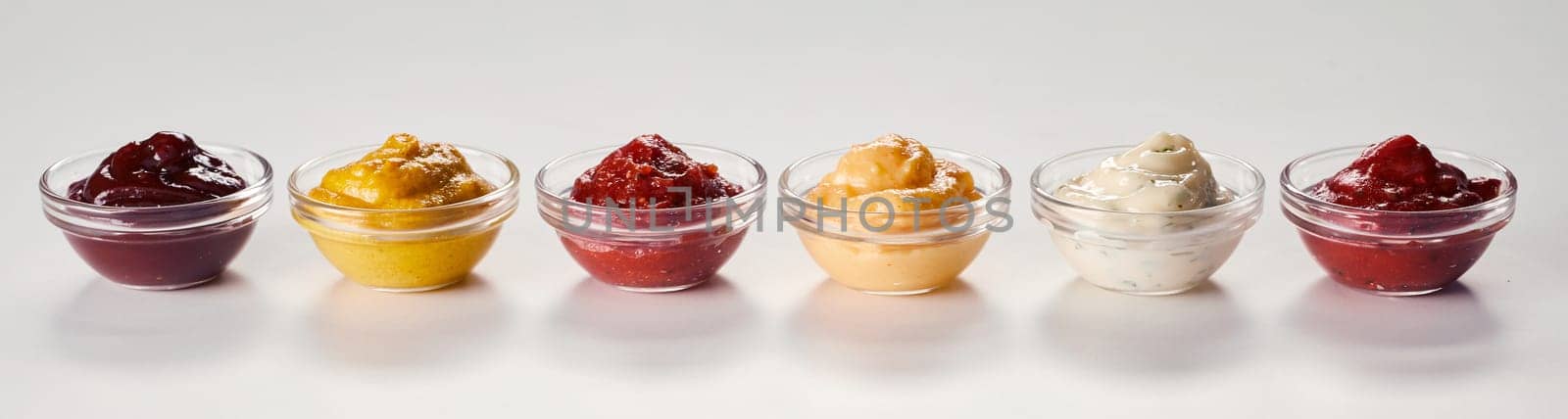 Small bowls of assorted sauces, dips, marinades and dressings on white with reflections in a panorama banner