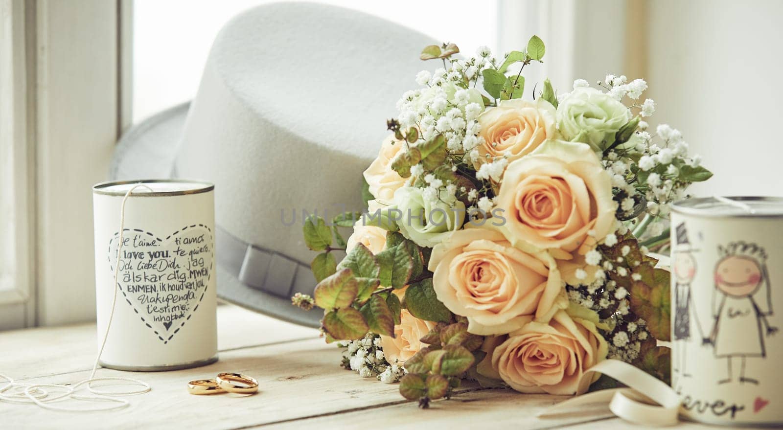 Marriage decor composition with brooms white hat, wedding rings, bridal bouquet of pink roses and pair of decorated cans with wishes, sitting on window sill