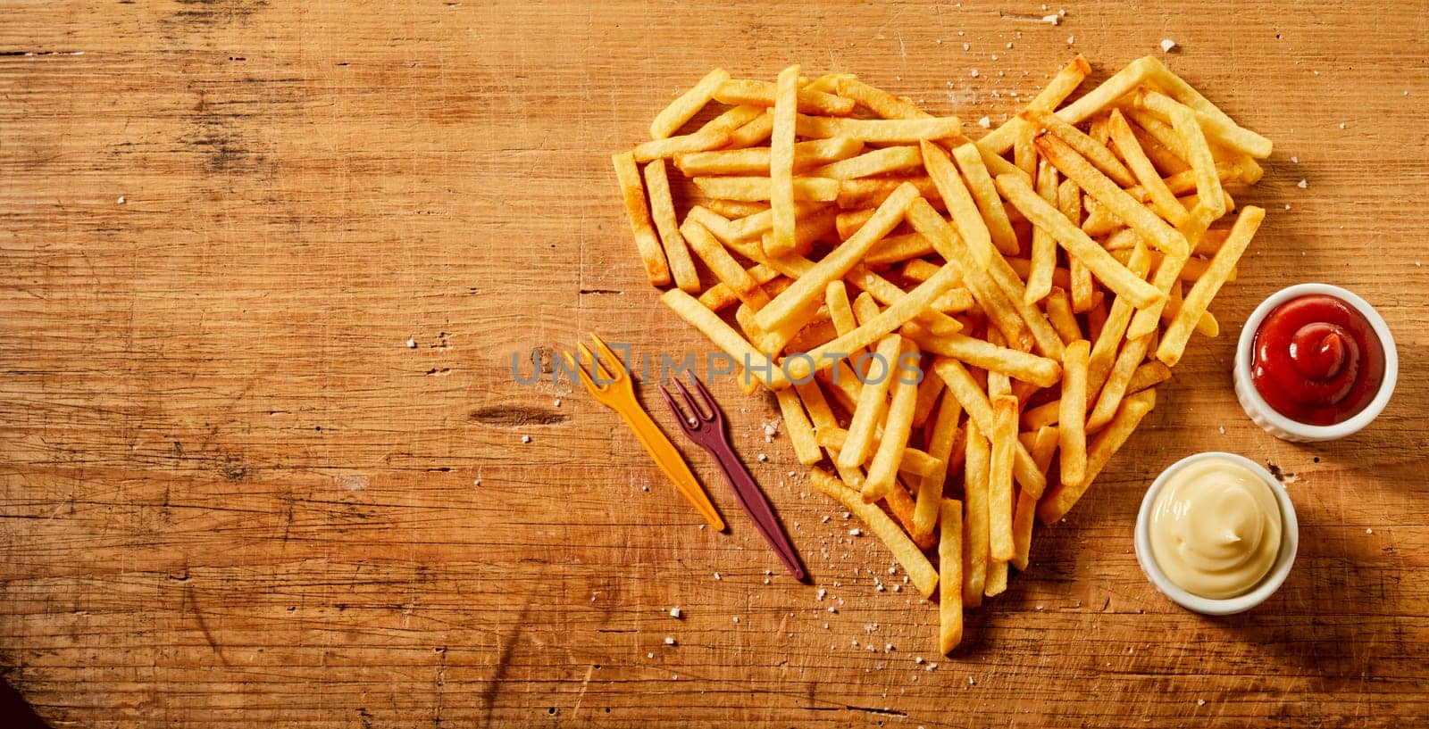 Heart shaped serving or fried potato chips, pommes frites or French fries on rustic wood with bowls of mustard and mayonnaise mix and tomato sauce viewed from overhead with copy space