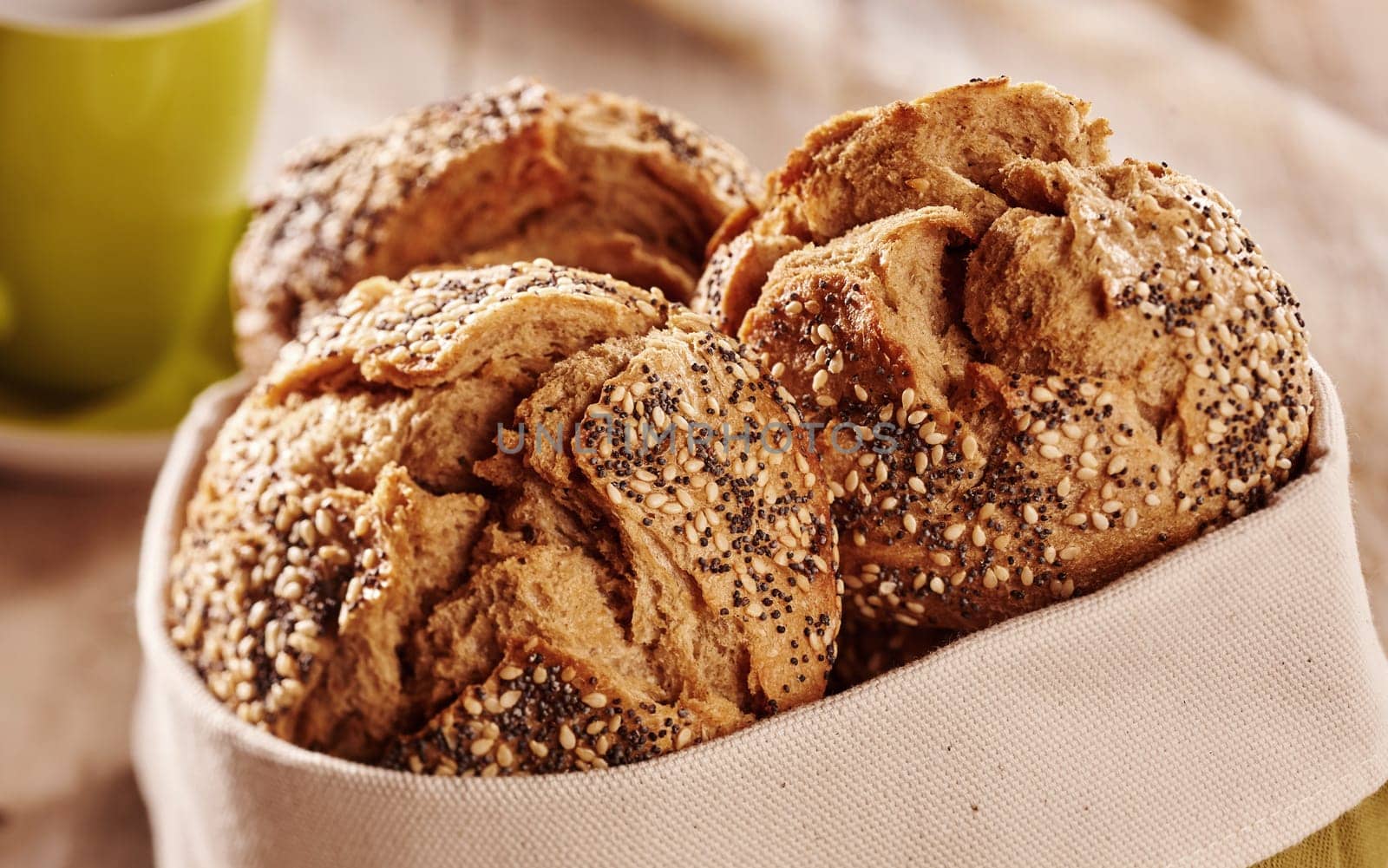Buns of fresh baked crunchy wholewheat bread with cracked crust topped with seeds, served in fabric bag with cup of beverage in background. Close-up view