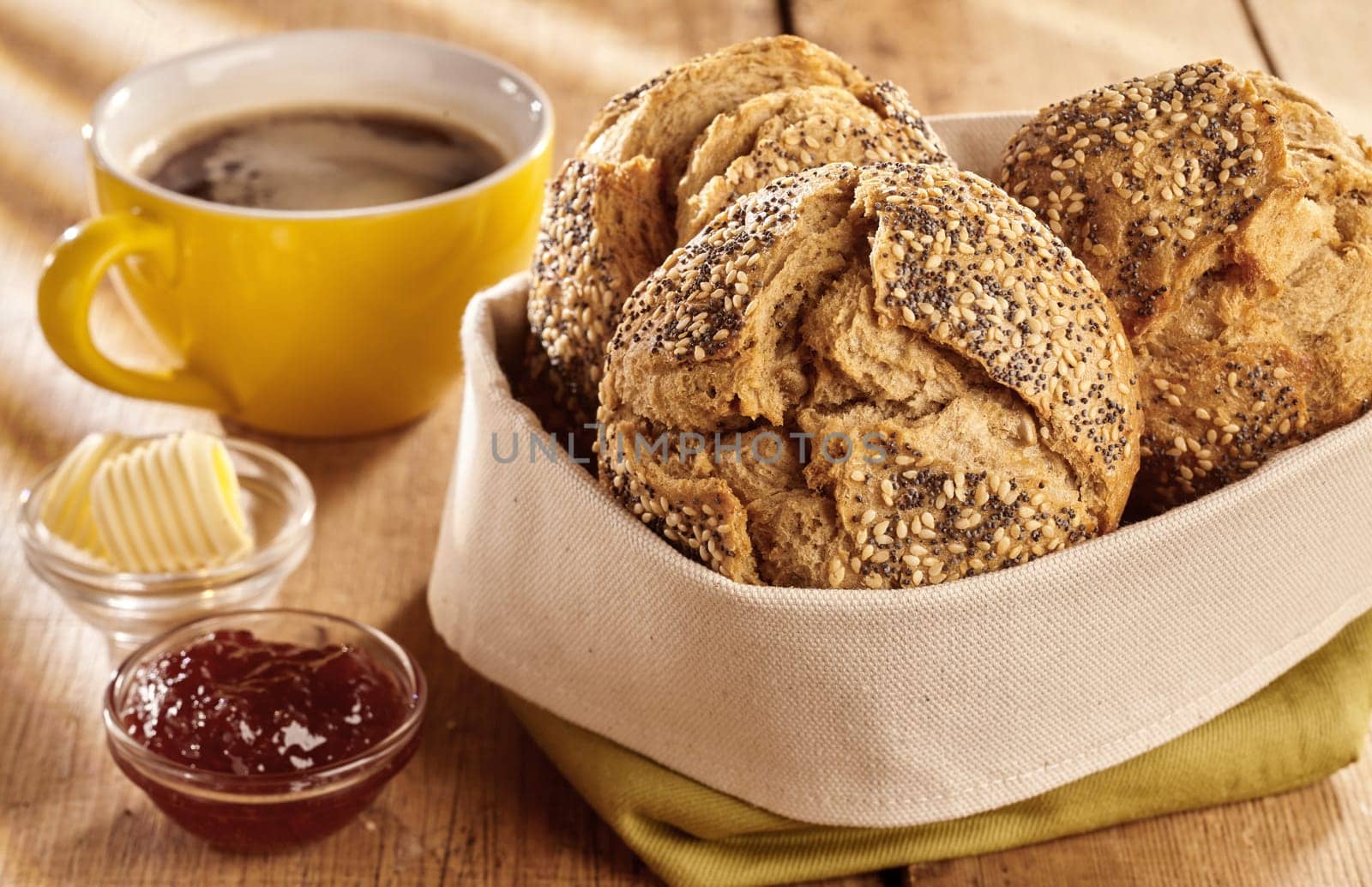 Fresh baked buns of brown wholewheat bread served for breakfast in fabric bag, with bowl of jam, curls of butter and cup of coffee, on wooden table and viewed in close-up
