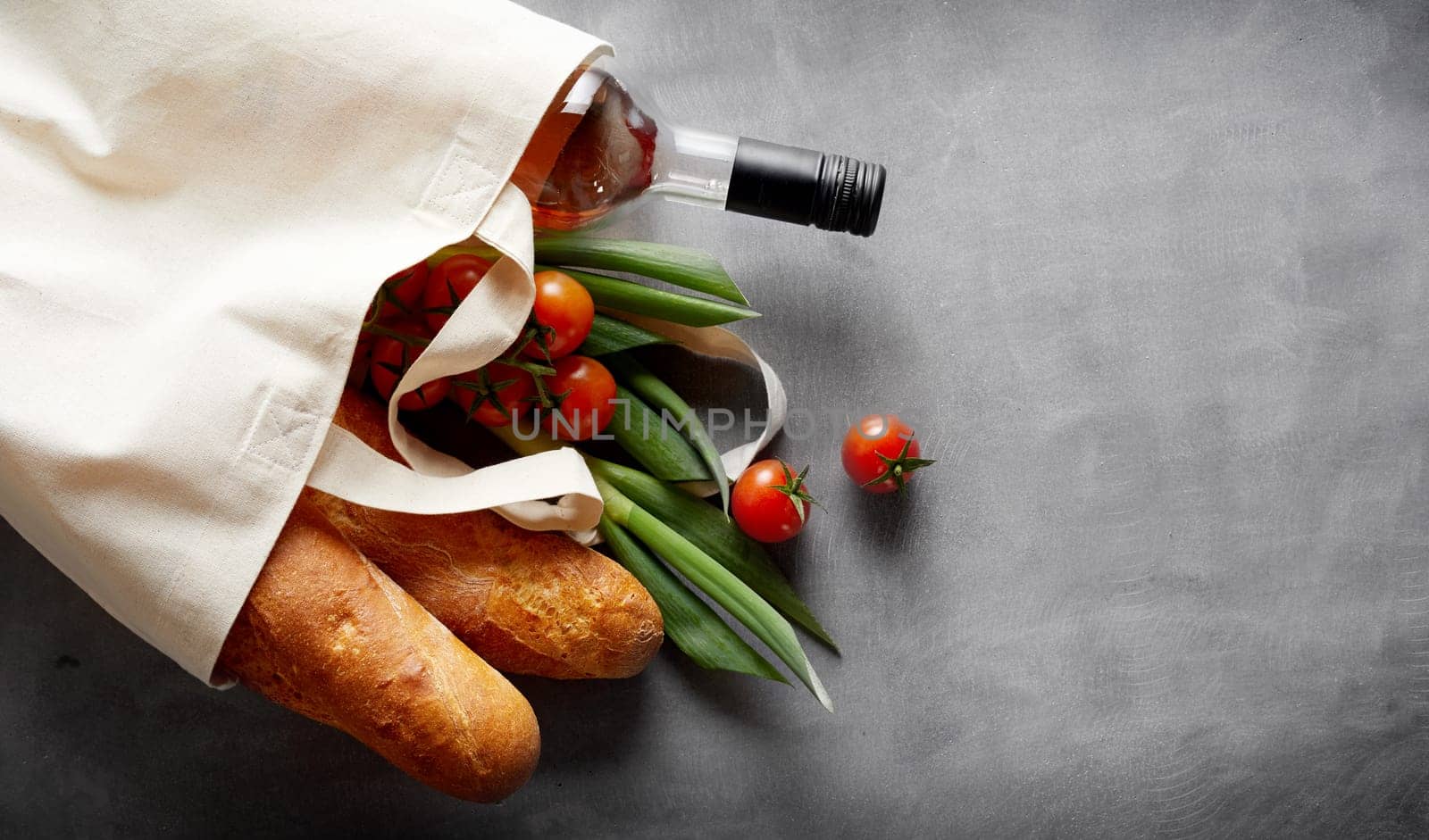Eco-friendly reusable textile bag with groceries and wine spilling out onto a slate background with copy space