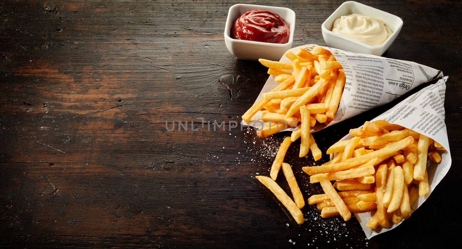Paper cones with salted French fries spilling out onto dark wood with copy space served with sauces
