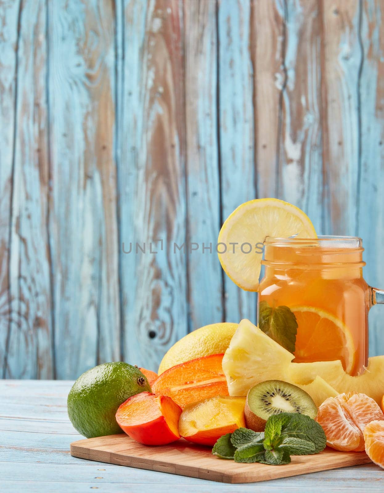 Fruit lemonade with ice, leaves of mint, lemon, pineapple, mango and lime slices standing on blue wooden table. Homemade lemonade with mix fruits. Healthy cool beverage, fruit juice