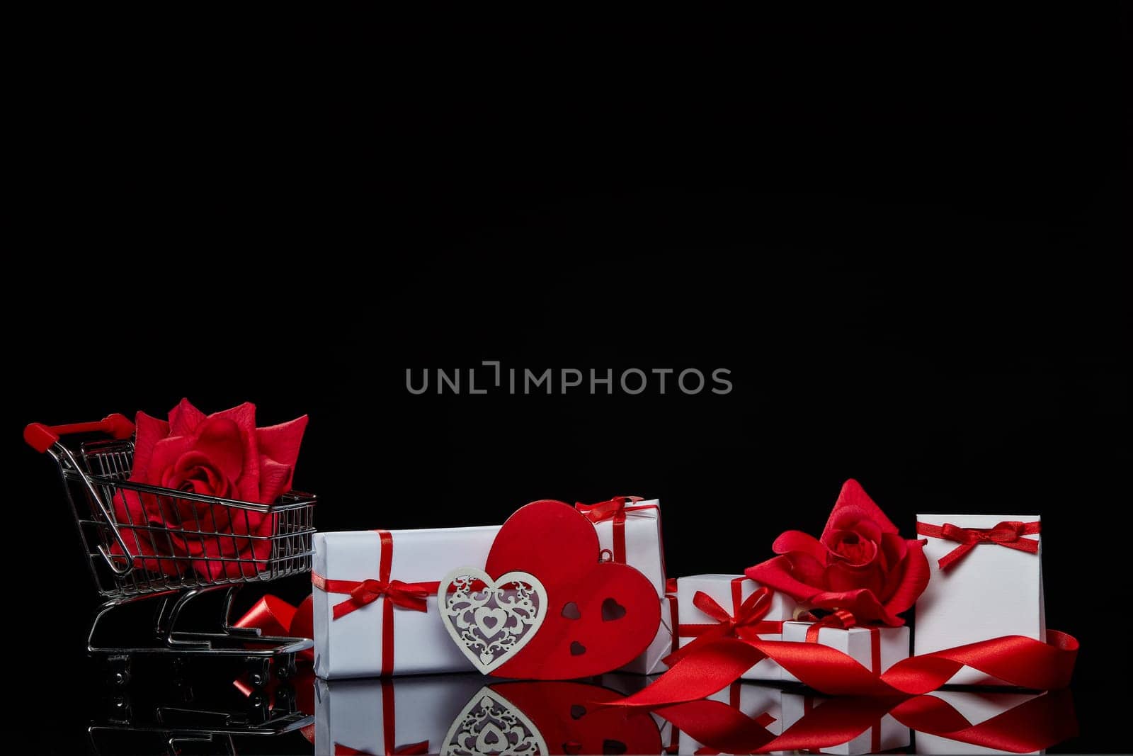 Shopping trolley with red roses, gift boxes and decorative heart on a black background with copy space. Valentine's day