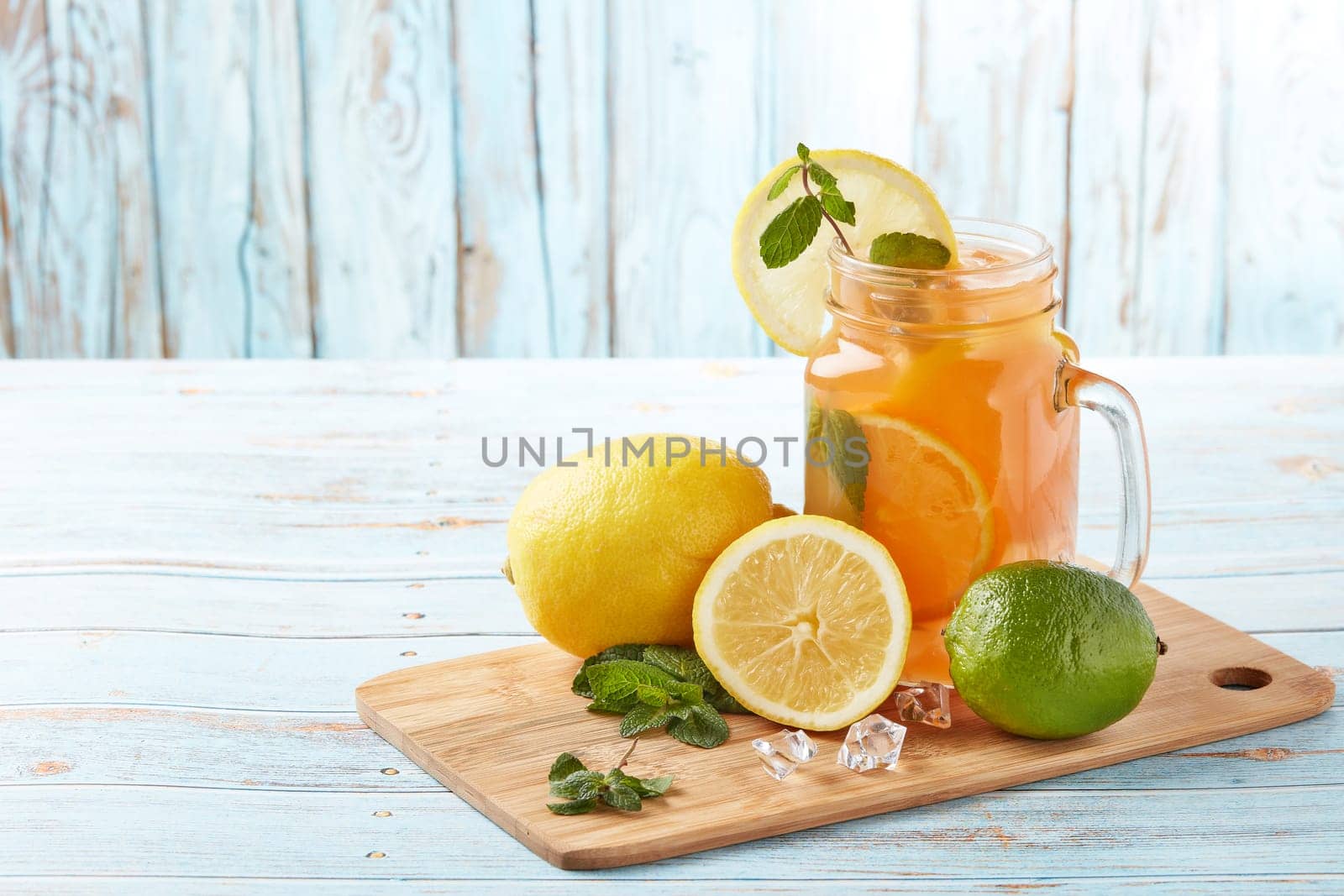 Citrus lemonade with ice, leaves of mint, lemon and lime slices standing on blue wooden table. Homemade lemonade with citruses. Healthy cool beverage