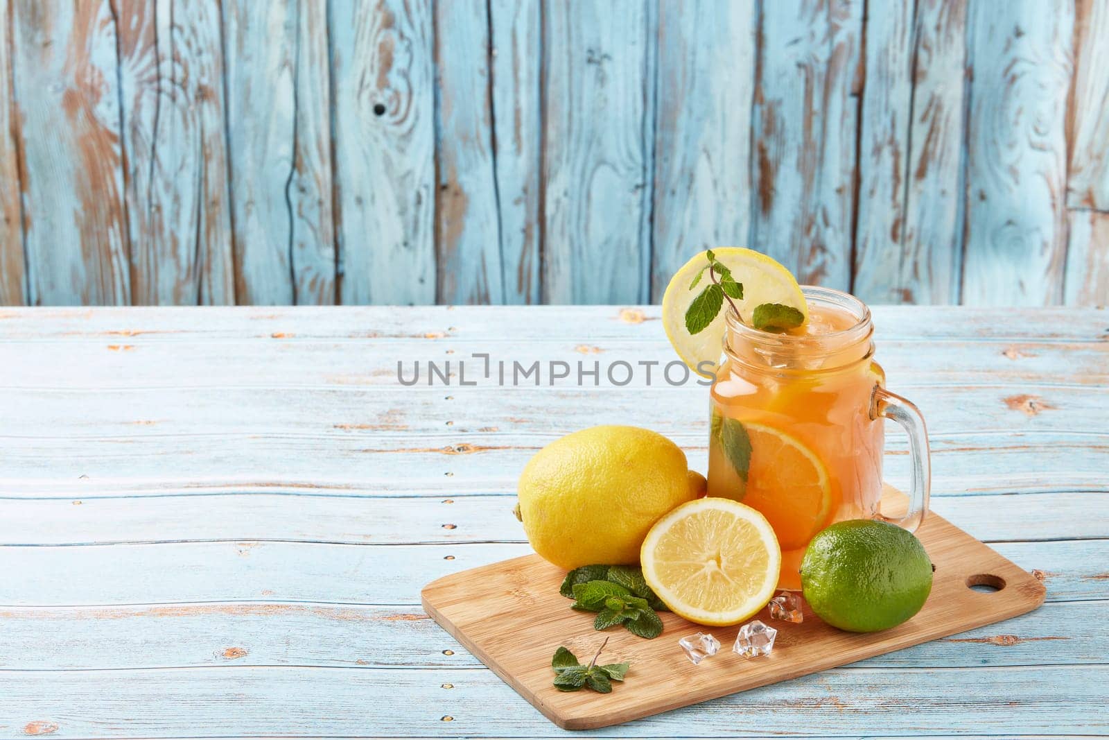 Citrus lemonade with ice, leaves of mint, lemon and lime slices standing on blue wooden table. Homemade lemonade with citruses. Healthy cool beverage