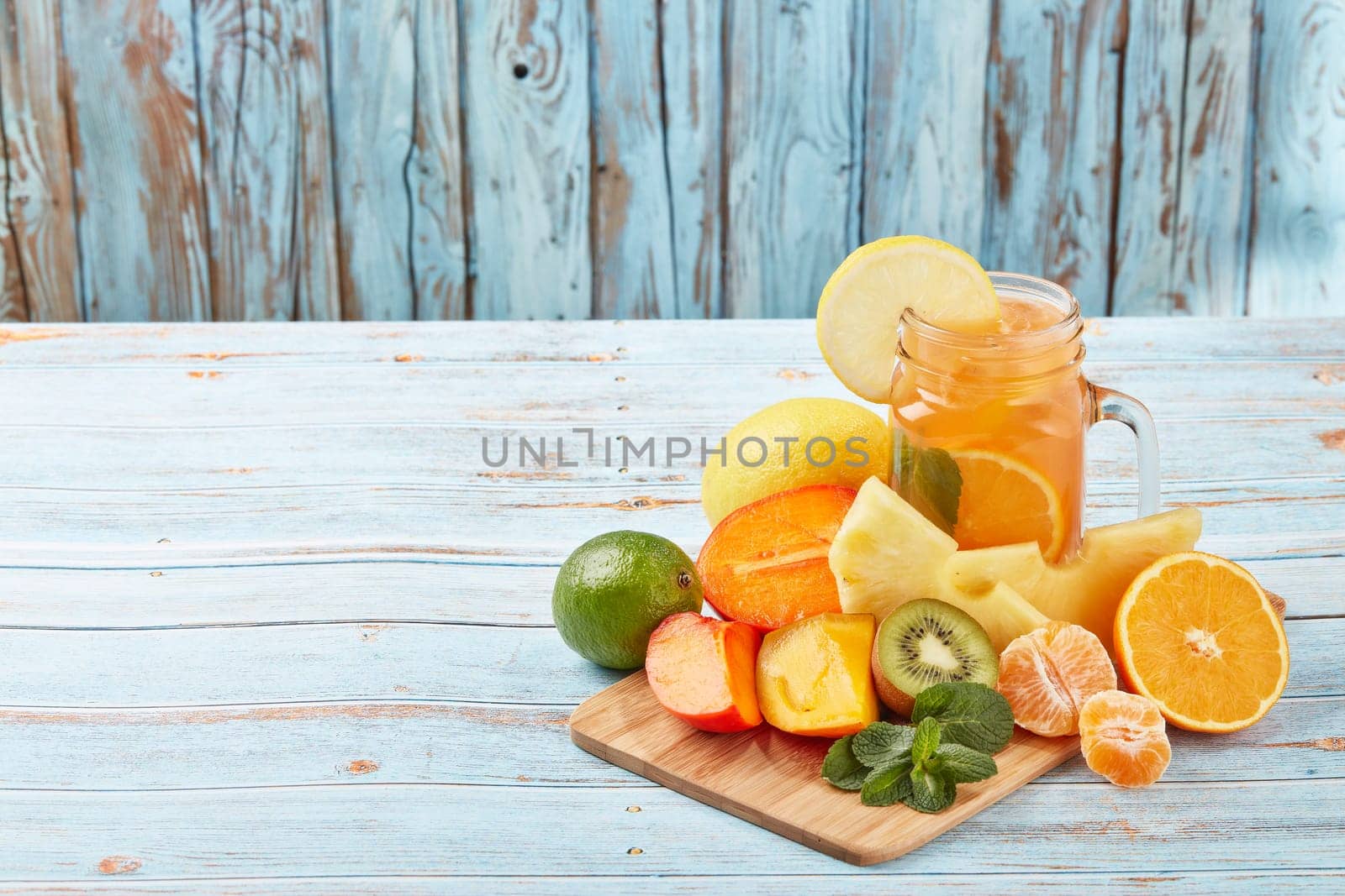 Fruit lemonade with ice, leaves of mint, lemon, pineapple, mango and lime slices standing on blue wooden table. Homemade lemonade with mix fruits. Healthy cool beverage, fruit juice